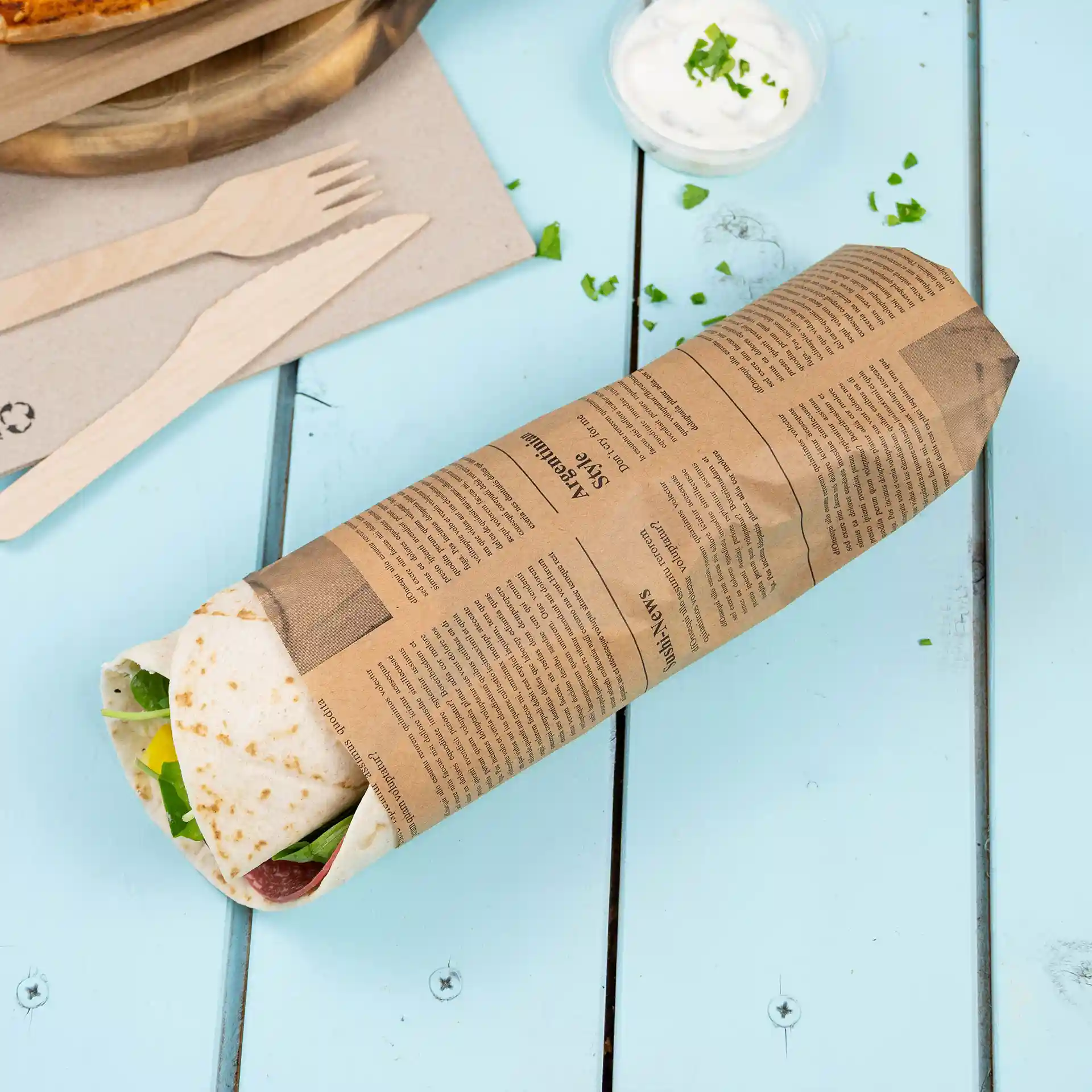 Article alimentaire emballé dans du papier kraft imprimé sur une surface en bois bleu clair, avec des ustensiles en bois et un petit bol de sauce à proximité.
