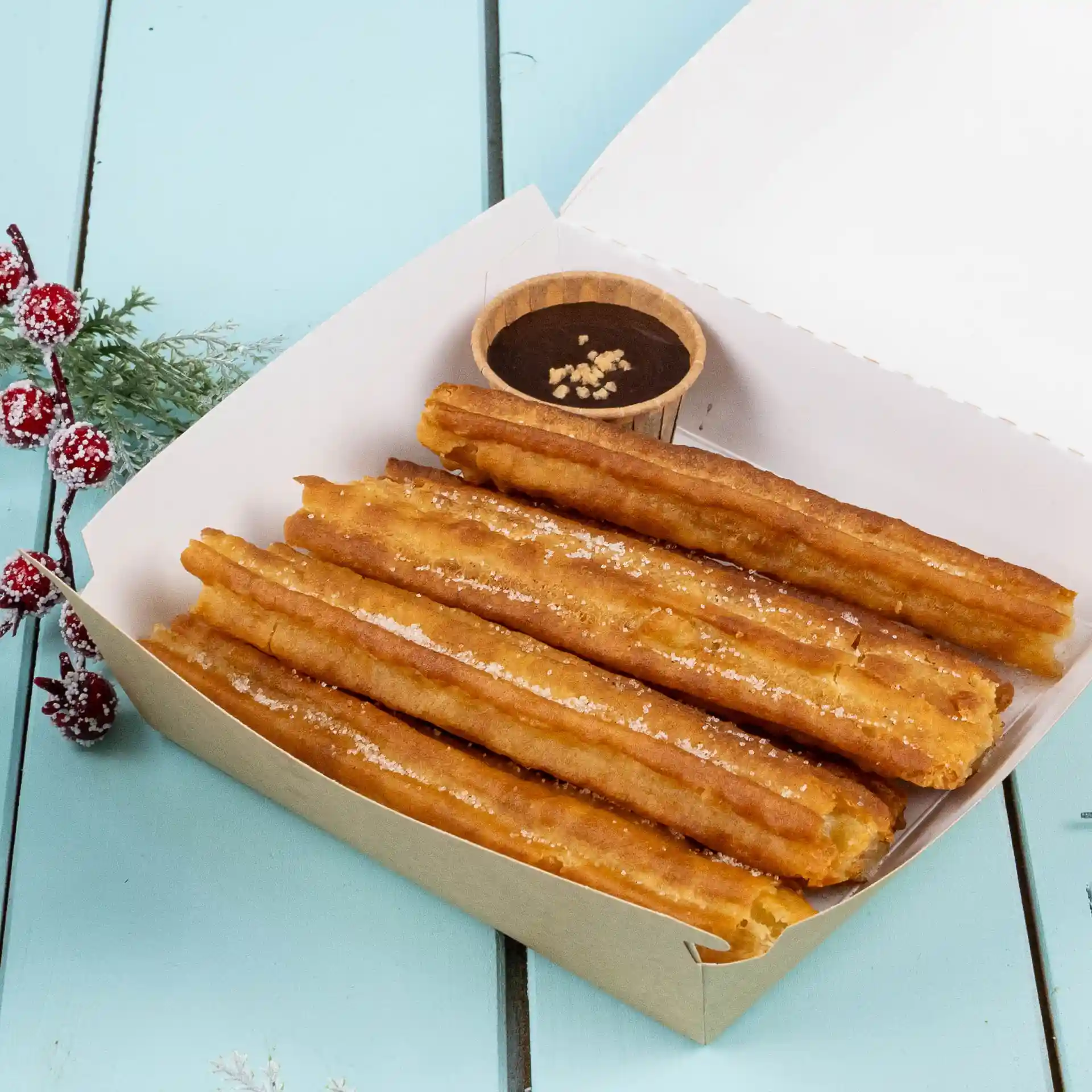 Churros in einer Papierbox mit einer kleinen braunen Tasse Schokoladensauce auf einer blauen Oberfläche.
