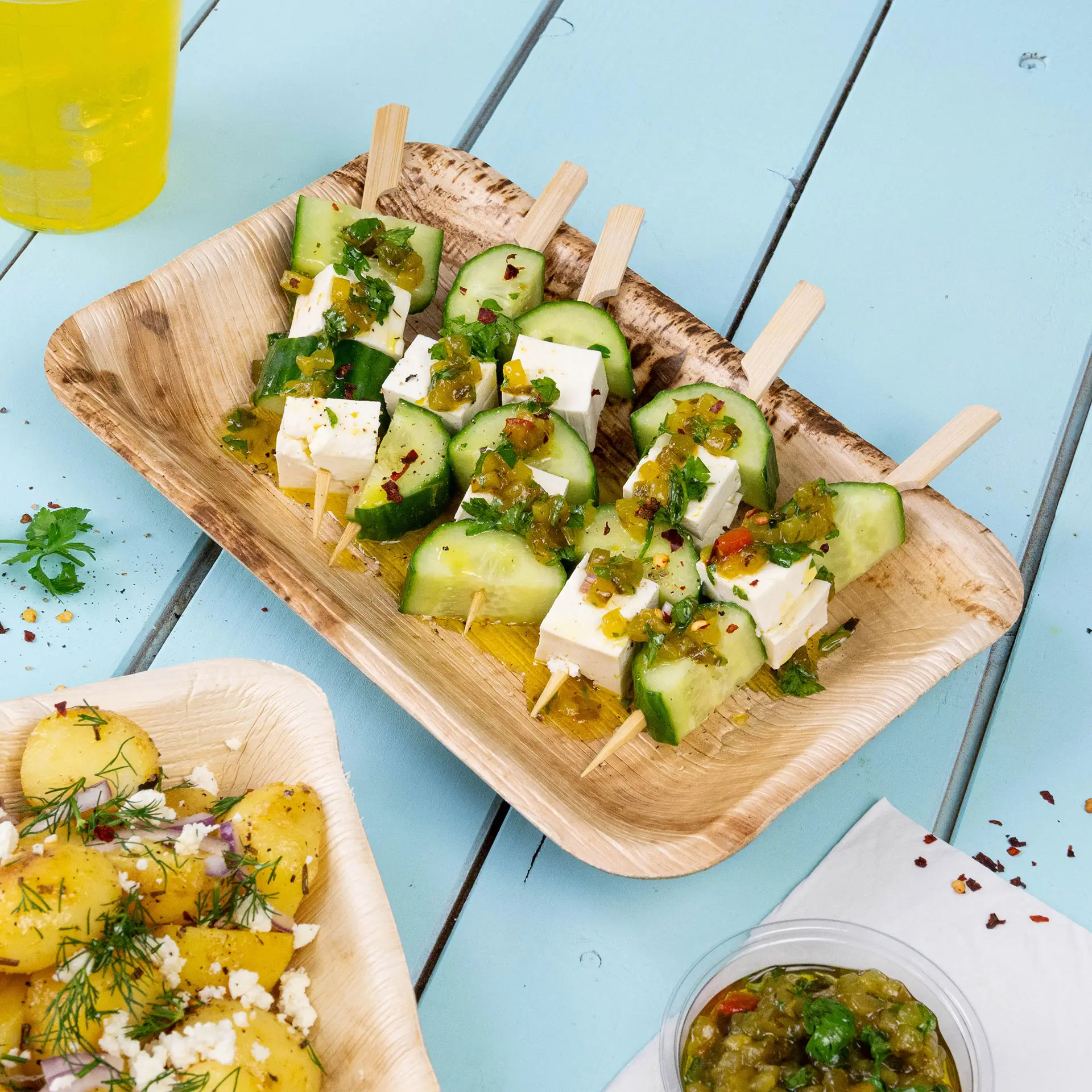 Un piatto rettangolare in foglia di palma con spiedini di cetriolo e feta, guarnito con salsa verde e coriandolo.