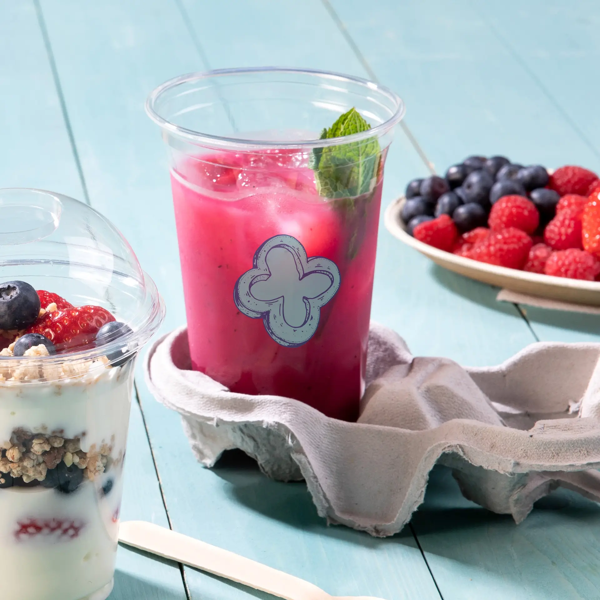 Bicchiere rPET trasparente da 20 oz con etichetta decorativa blu, riempito di una bevanda rosa e guarnito con menta, su uno sfondo di stoffa.