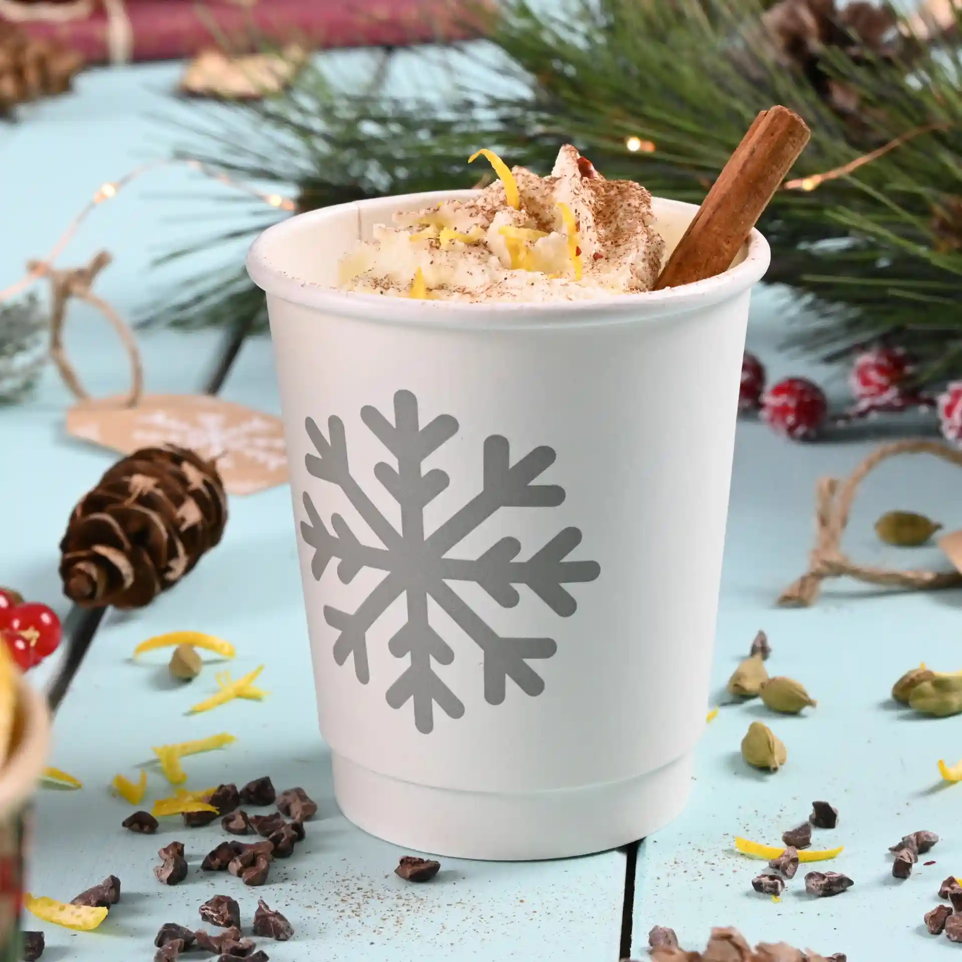 Bicchiere bianco con design di fiocco di neve grigio, riempito di panna montata e un bastoncino di cannella, su una superficie blu.