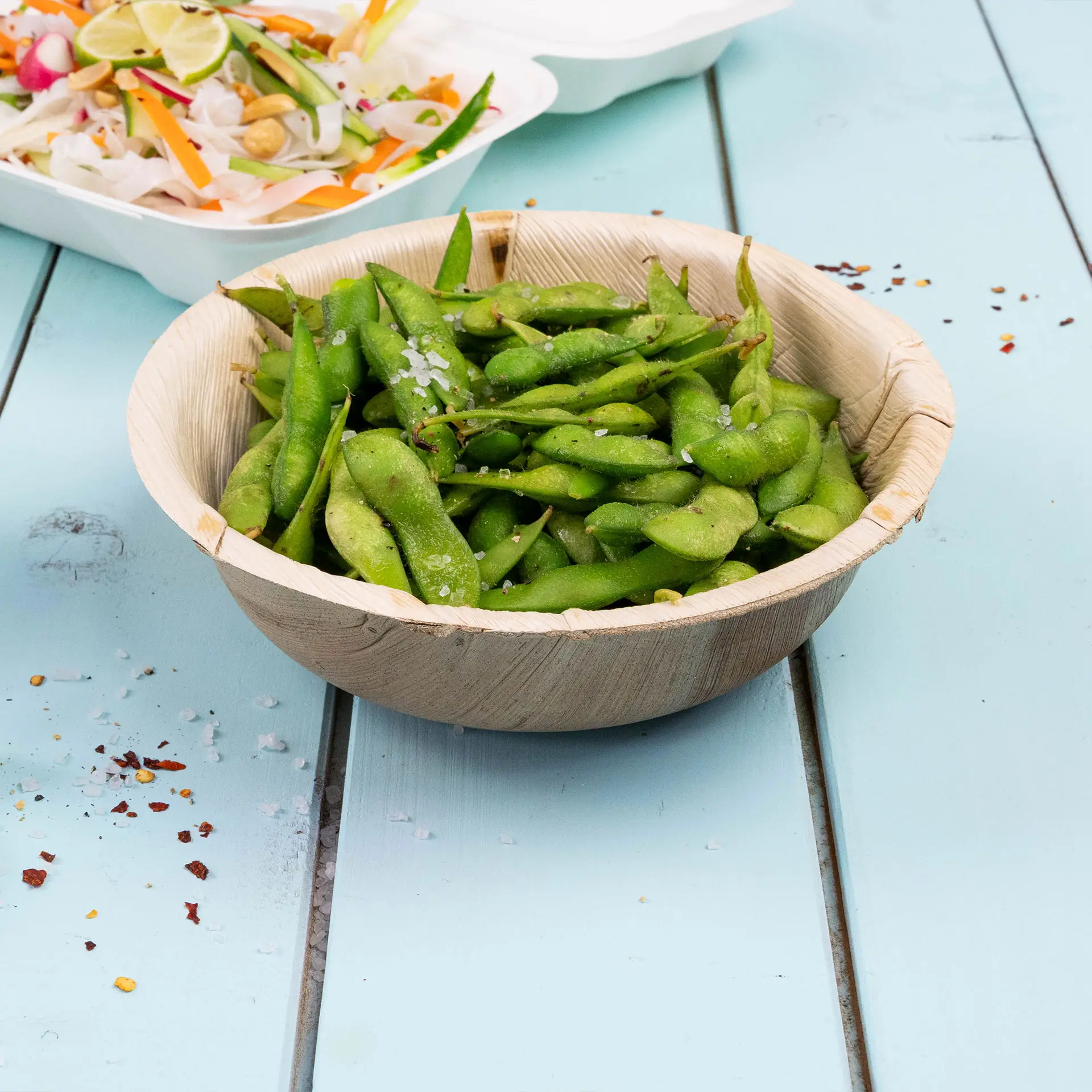 Ciotola rotonda in foglia di palma riempita di edamame verdi, cosparsa di sale, su una superficie in legno blu.