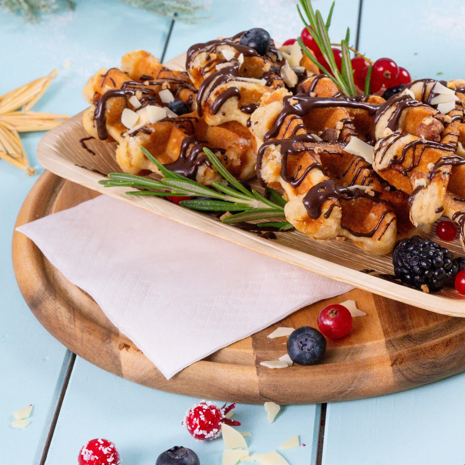 Un vassoio di legno tiene waffle guarniti con cioccolato, frutti di bosco e rosmarino, accanto a un tovagliolo bianco.