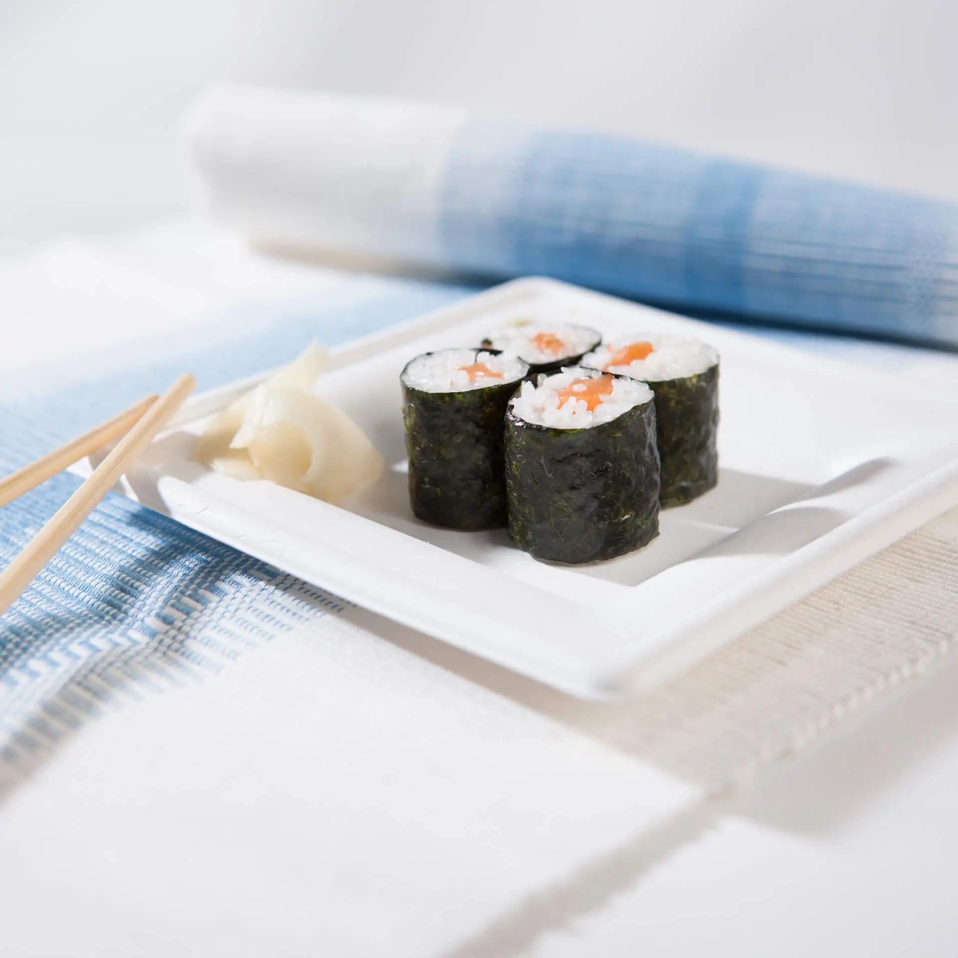 Quadratische Zuckerrohrplatte mit Sushi-Rollen, eingelegtem Ingwer und Essstäbchen auf einem strukturierten Stoffhintergrund.