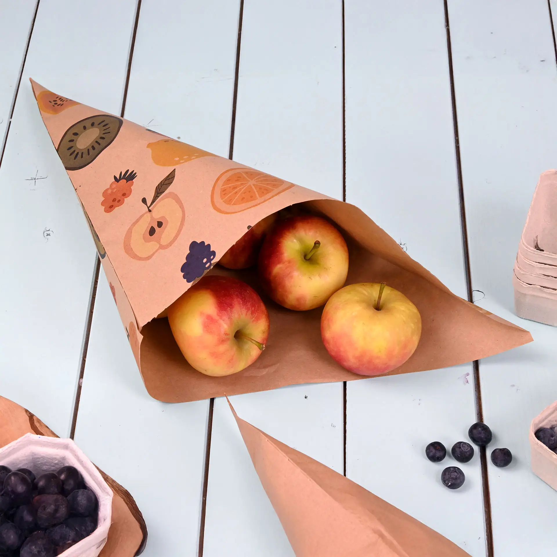 Brauner Papiertrichter mit Obstmotiv, enthält drei rote Äpfel, auf einer hellblauen Tischoberfläche.