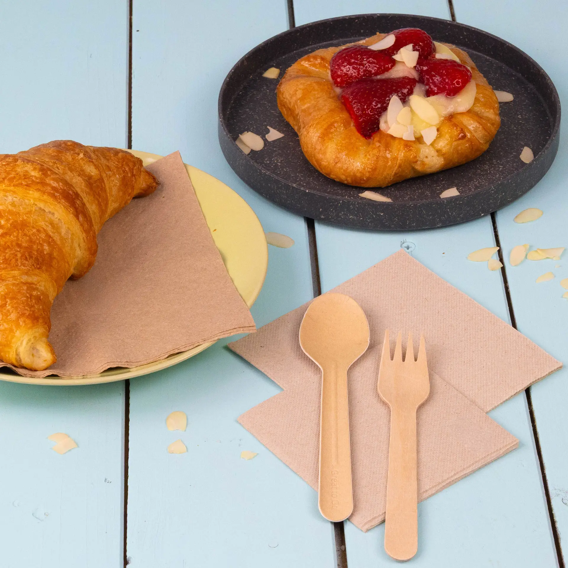 Image d'un croissant sur une assiette jaune, d'une pâtisserie aux fruits sur une assiette noire et de serviettes recyclées avec une cuillère et une fourchette.