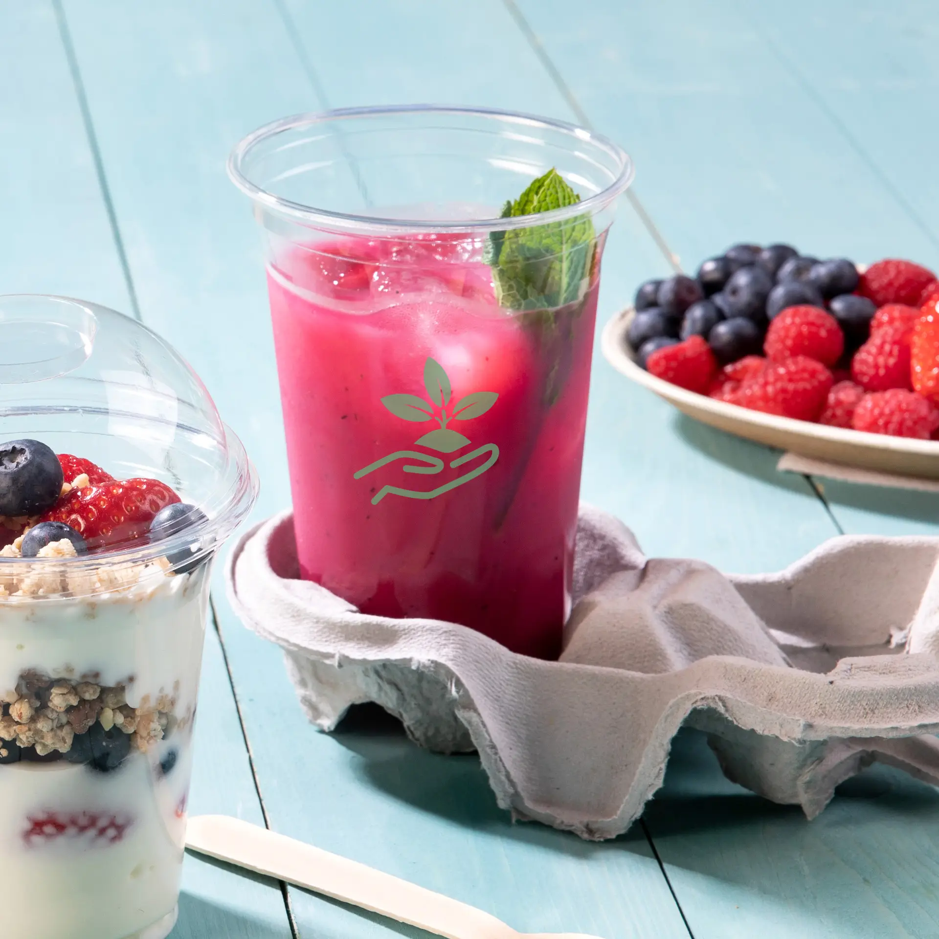 Un bicchiere trasparente in rPET riempito di una bevanda rosa e una foglia di menta; un cartone per uova regge il bicchiere; un parfait di yogurt e bacche sono nei dintorni.