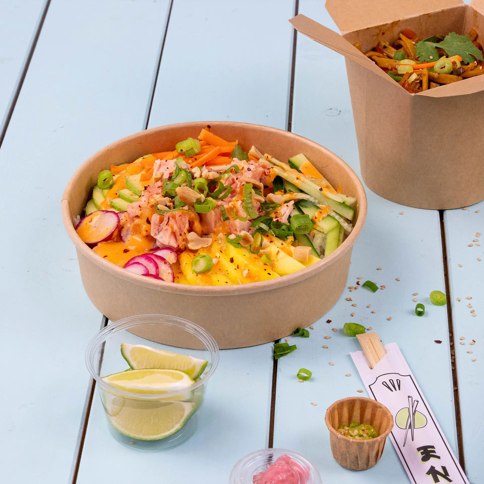 Braune Papierschüssel mit gemischtem Gemüse und Toppings, Limettenscheiben und einem zweiten Behälter im Hintergrund.