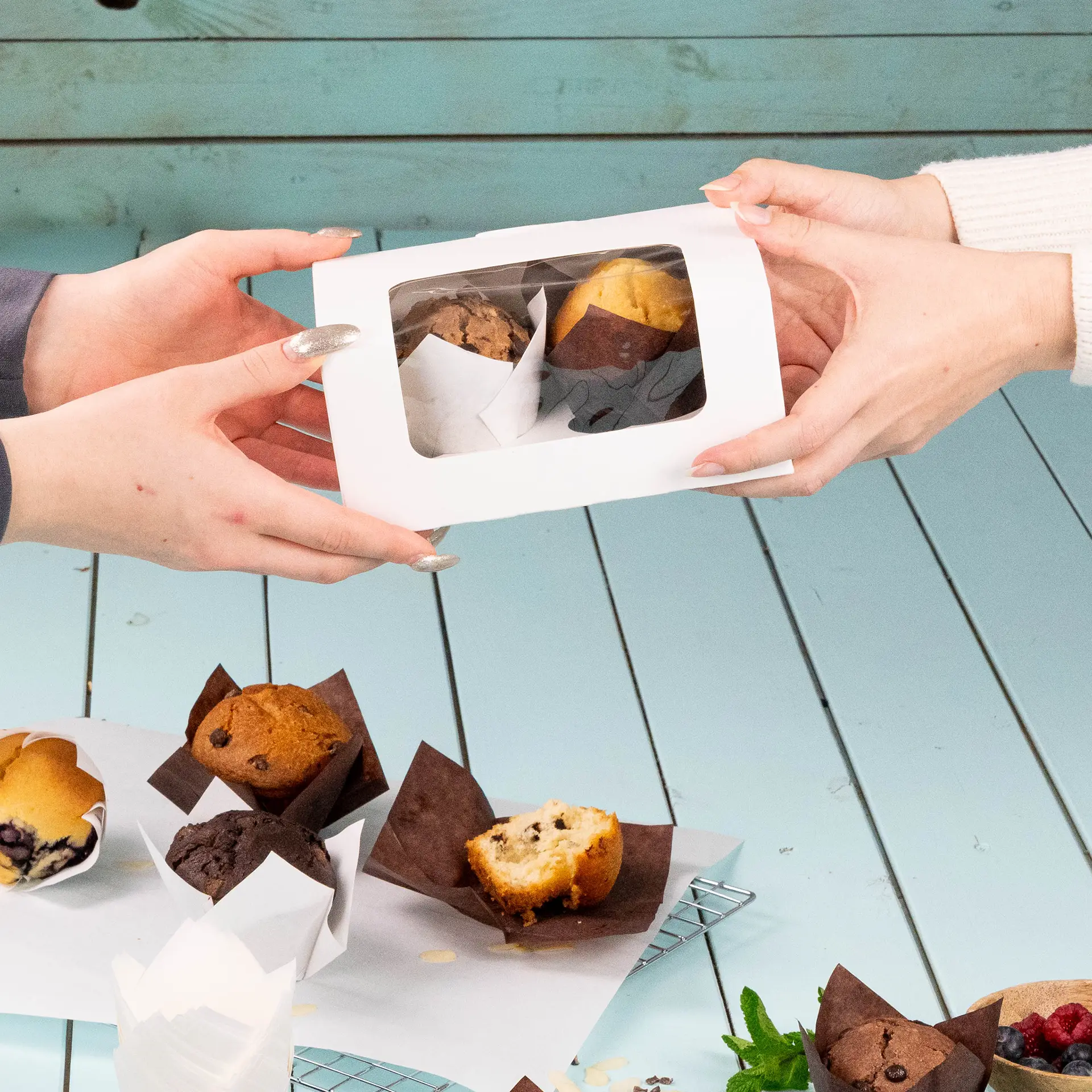 Zwei Hände tauschen eine weiße Box mit klarem Fenster aus, die zwei Muffins enthält, mit mehreren Muffins auf einem Tisch.