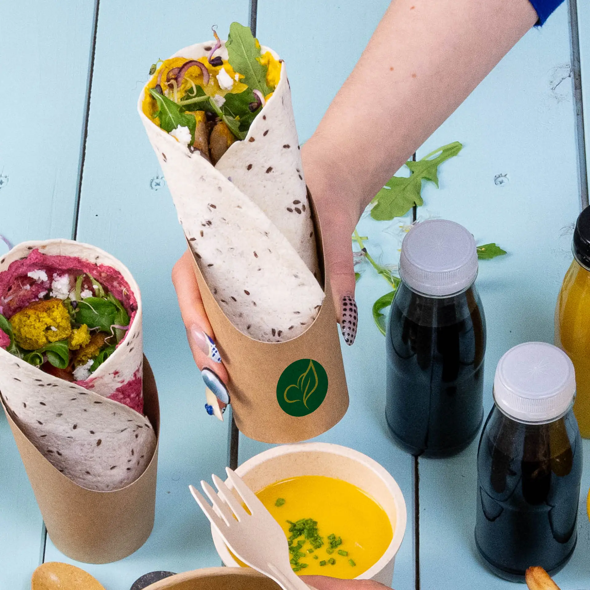 Un gobelet wrap imprimé personnalisé marron contenant un wrap rempli ; un autre gobelet wrap à côté. Plusieurs bouteilles et un bol avec de la sauce sont à proximité.