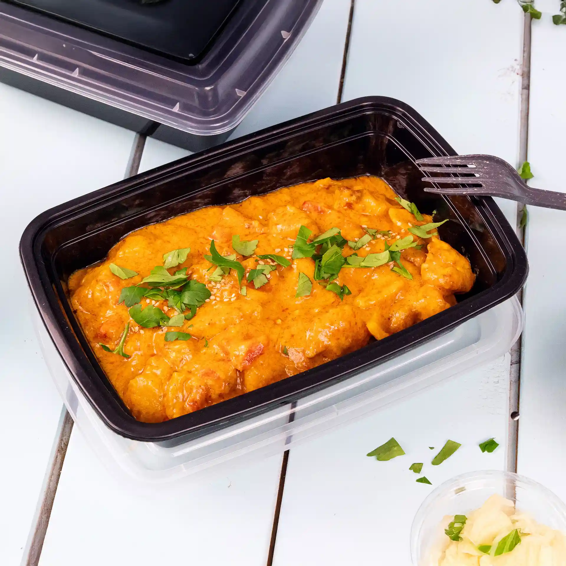 Contenitore rettangolare nero riutilizzabile con coperchio, contenente cibo arancione guarnito con erbe verdi e semi di sesamo.