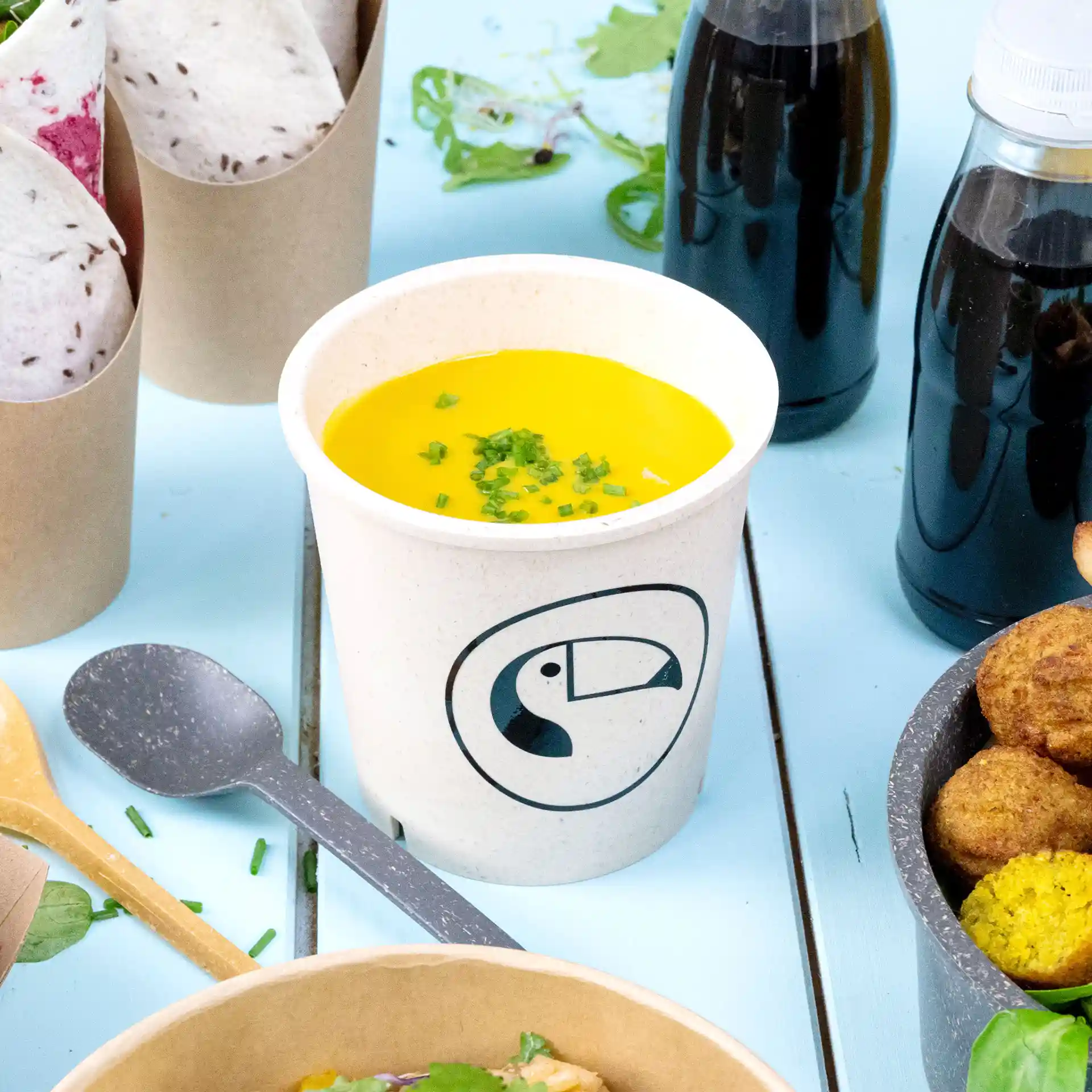 Runde cremeweiße Suppenschale mit Logo, gefüllt mit gelber Suppe und garniert mit gehacktem Schnittlauch, auf blauer Fläche.