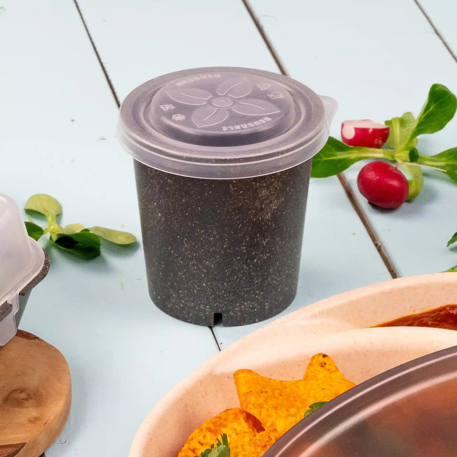 Bicchiere rotondo trasparente con coperchio, posato su una superficie azzurra, circondato da foglie verdi e tortilla chips.