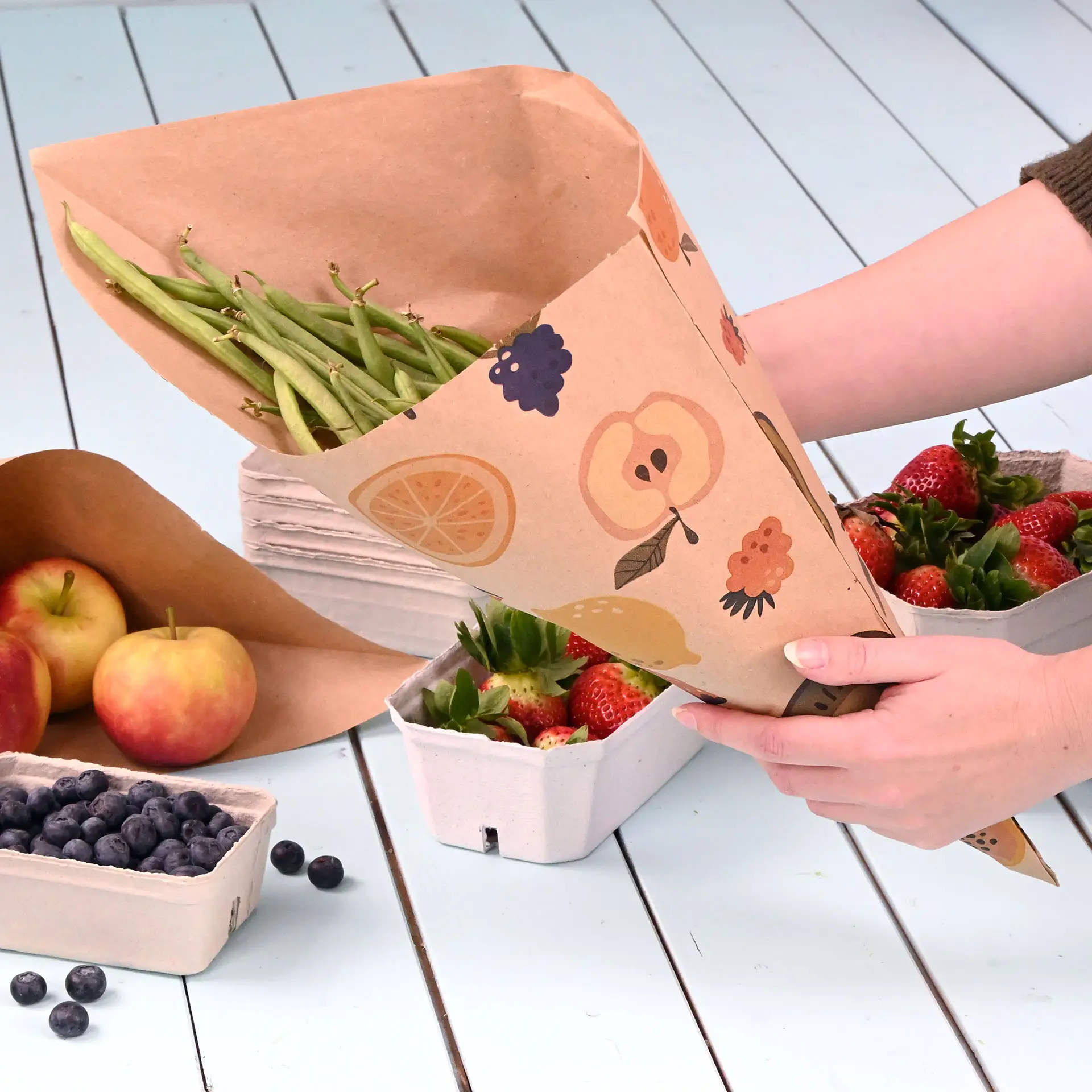 Eine Hand hält einen braunen Papiertütencone mit grünen Bohnen, umgeben von Erdbeeren, Äpfeln und Heidelbeeren auf einer hellblauen Oberfläche.