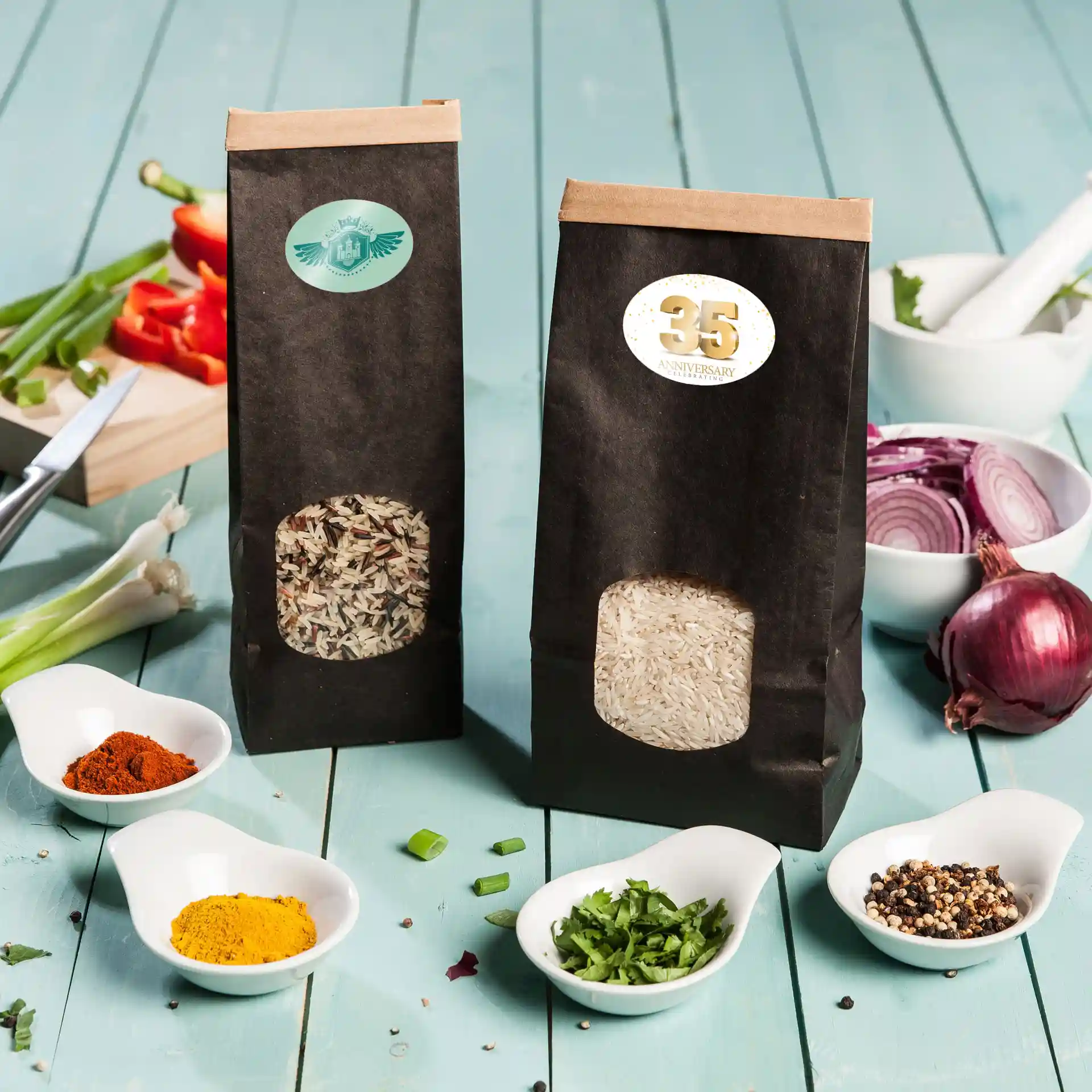 Deux sacs en papier noir contenant du riz, avec des étiquettes visibles, entourés de diverses épices et légumes hachés sur une surface en bois bleu.