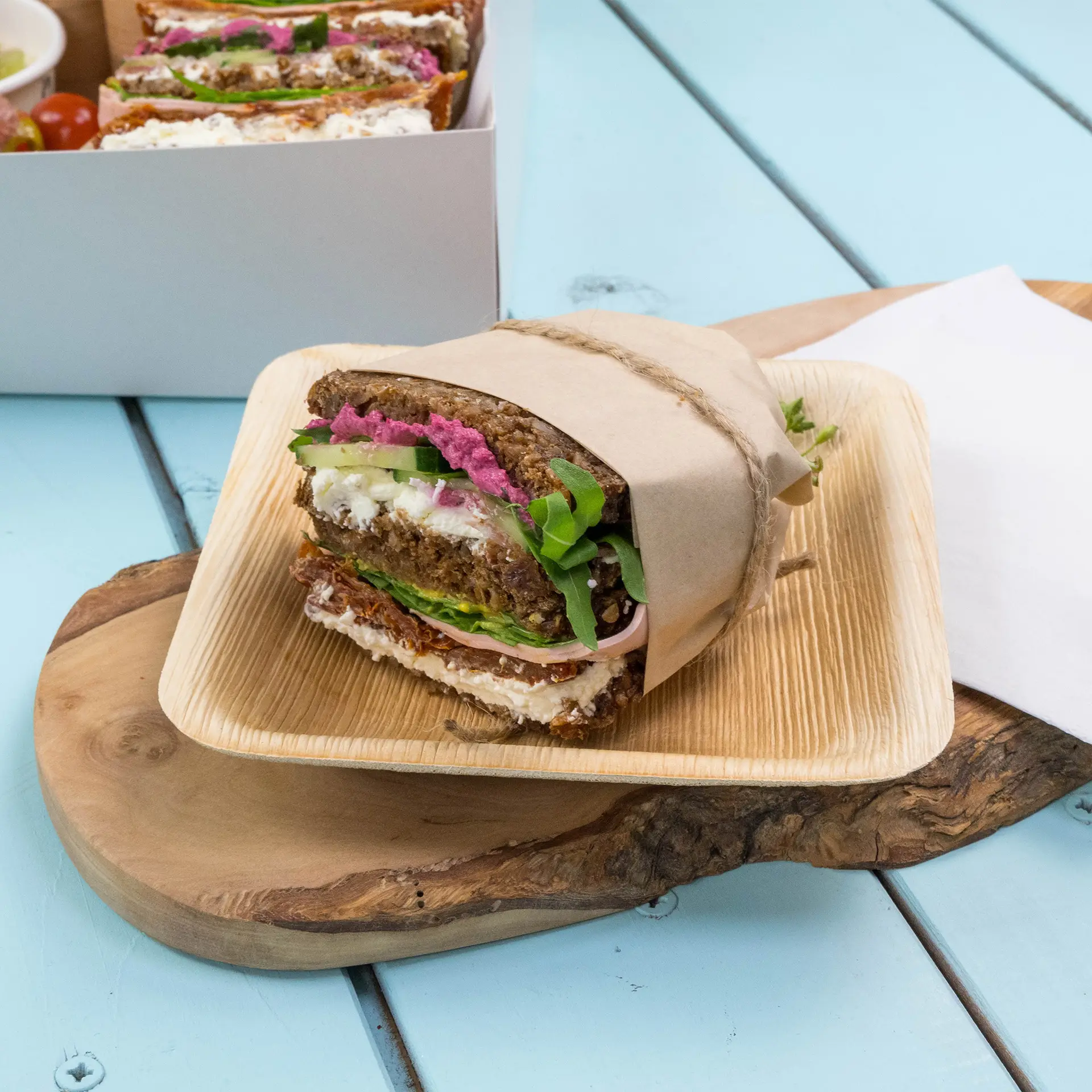 Braunes Geschenkpapier mit Sandwich, gefüllt mit Schichten von Gemüse, Frischkäse und rosa Aufstrich auf einem Holzbrett.