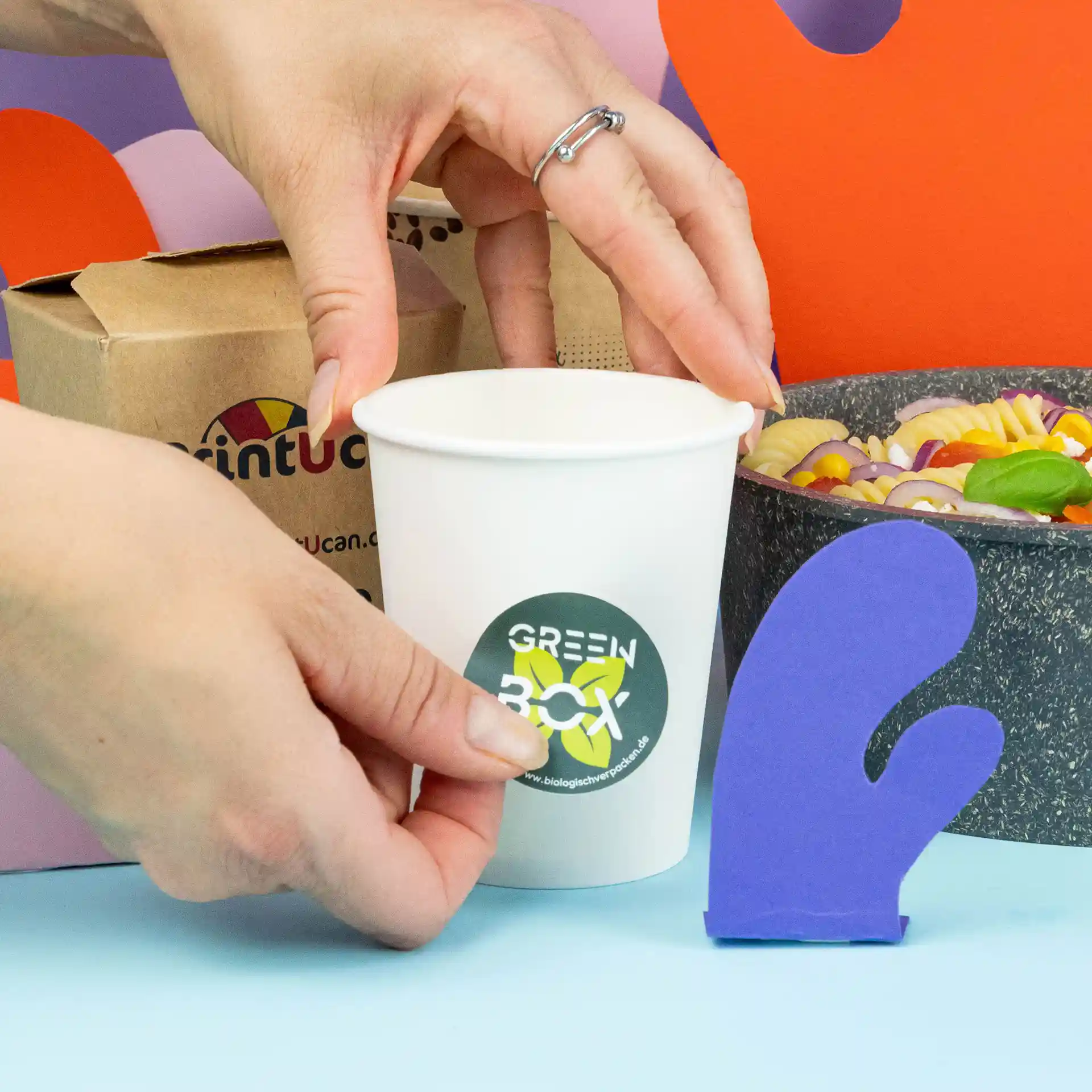 Une main place une étiquette ronde sur une tasse blanche, avec un gant de cuisine violet et un bol de pâtes en arrière-plan.