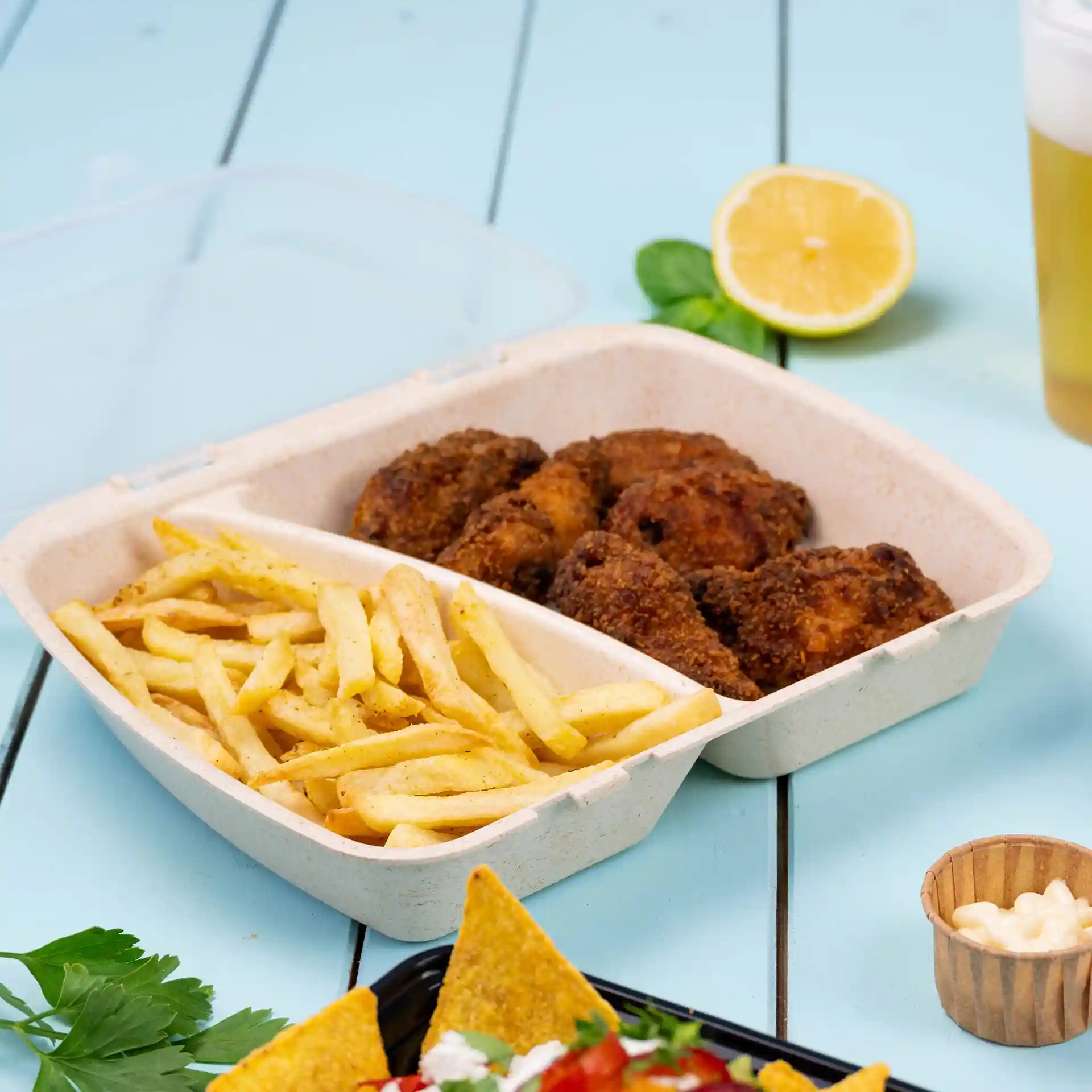 Behälter mit zwei Fächern, gefüllt mit gebratenem Hühnchen und Pommes, auf einer blauen Oberfläche mit Zitrone und Getränk.