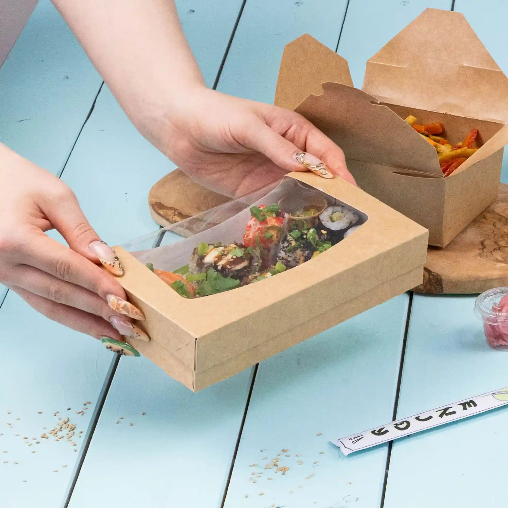 Boîte en carton avec sushi et salade, avec un couvercle transparent, sur une surface bleue.