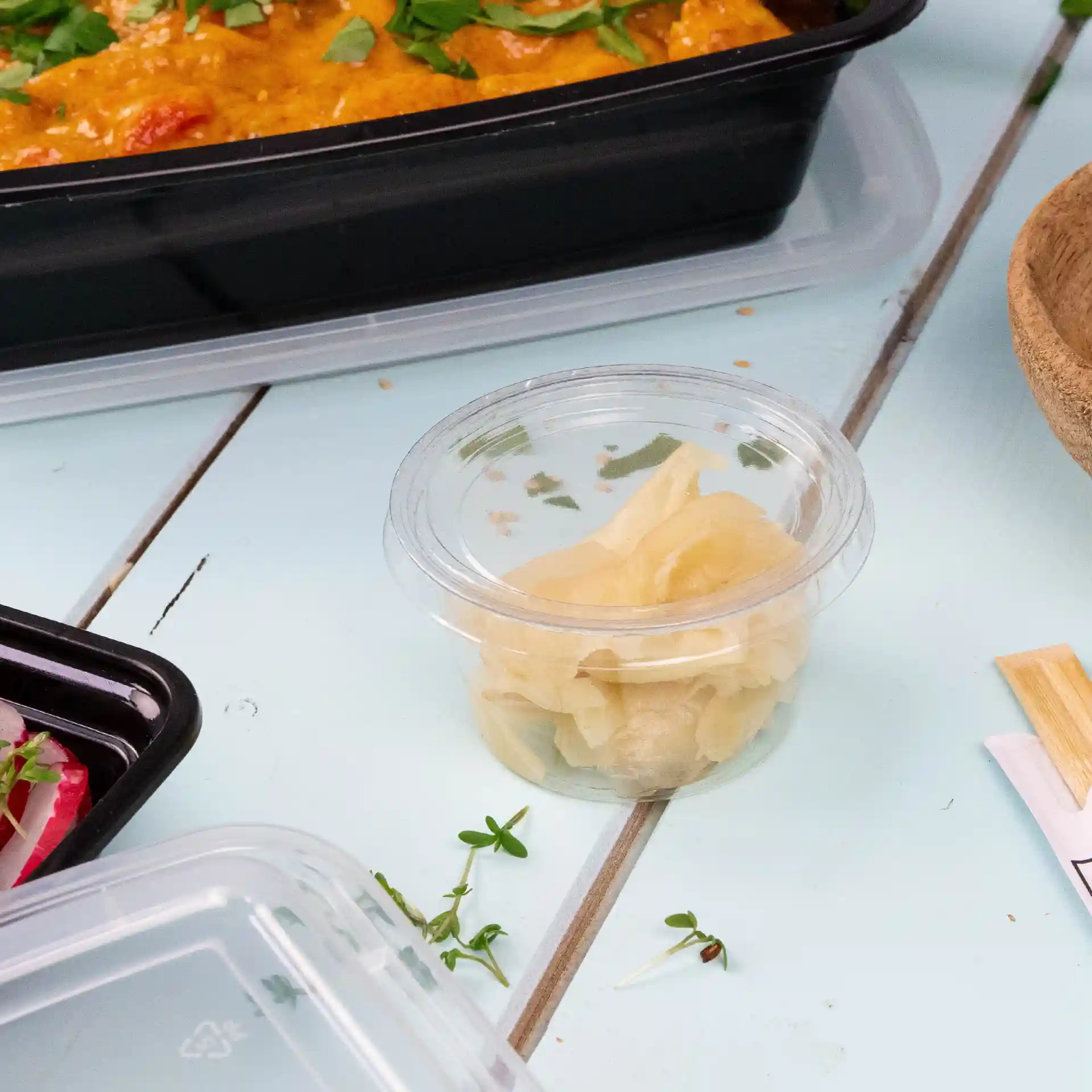 Klare PLA-Behälter mit eingelegtem Ingwer, auf einer hellblauen Oberfläche mit anderen Lebensmitteln in der Nähe.