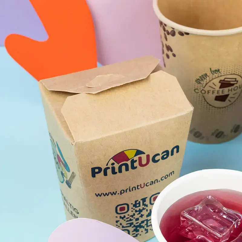 Contenants en carton brun pour nouilles ramen avec logo, placés sur une surface bleue avec divers garnitures et des baguettes.