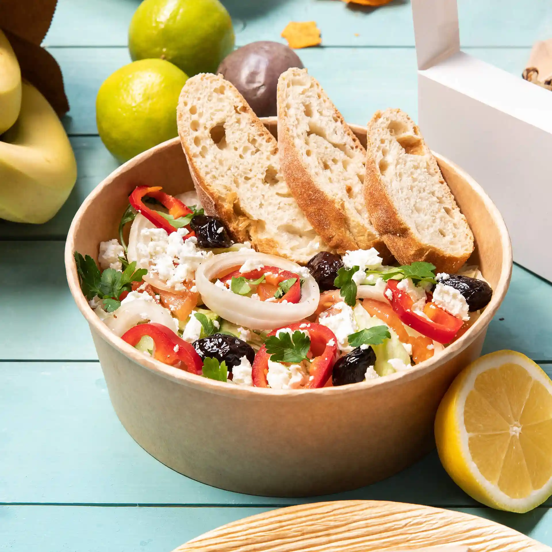 Ciotola rotonda in cartone marrone riempita di insalata, guarnita con fette di pane e circondata da frutta su una superficie blu.