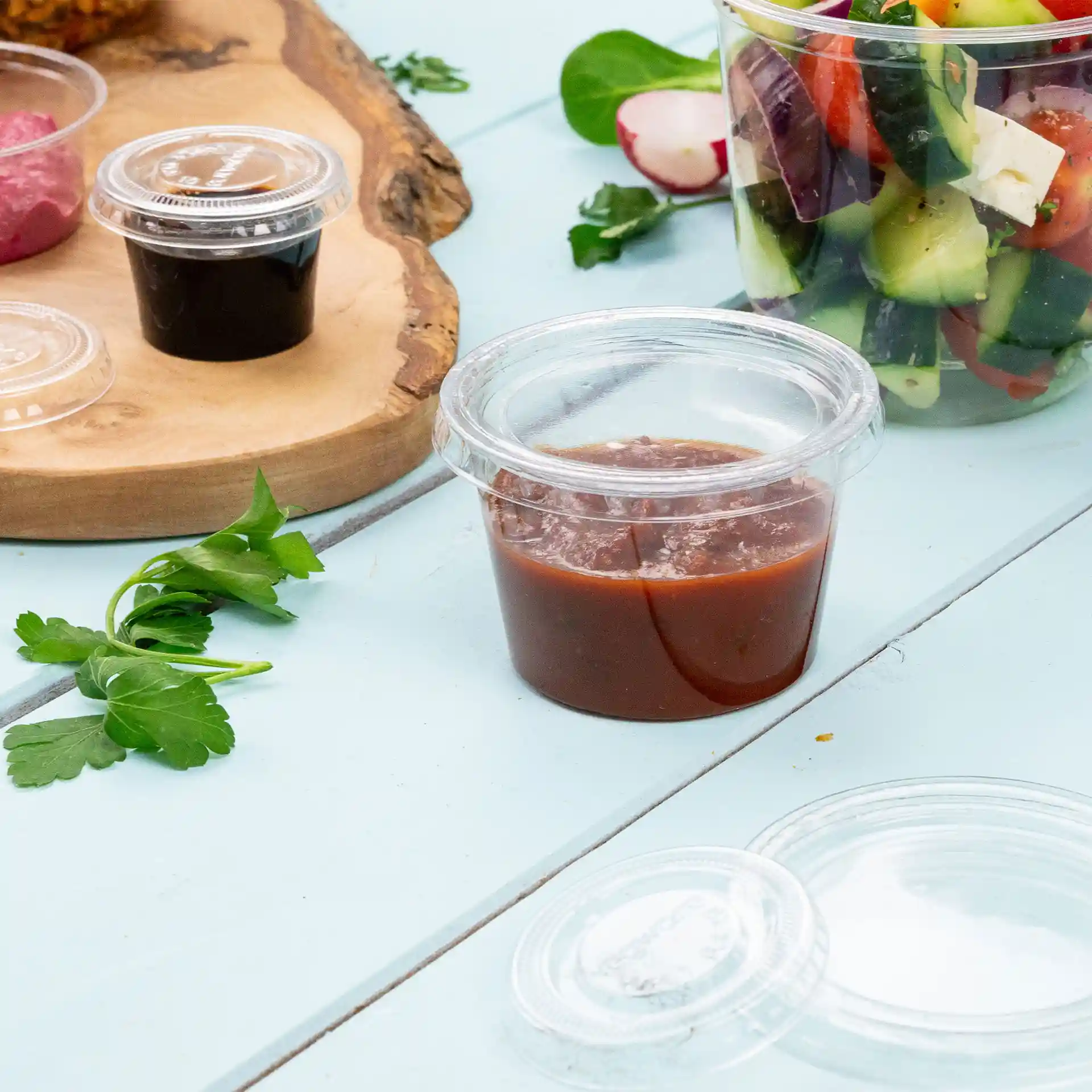 Bicchiere rotondo trasparente con salsa, capacità di 100 ml, posizionato su una superficie azzurro chiaro con prezzemolo e altri alimenti vicini.