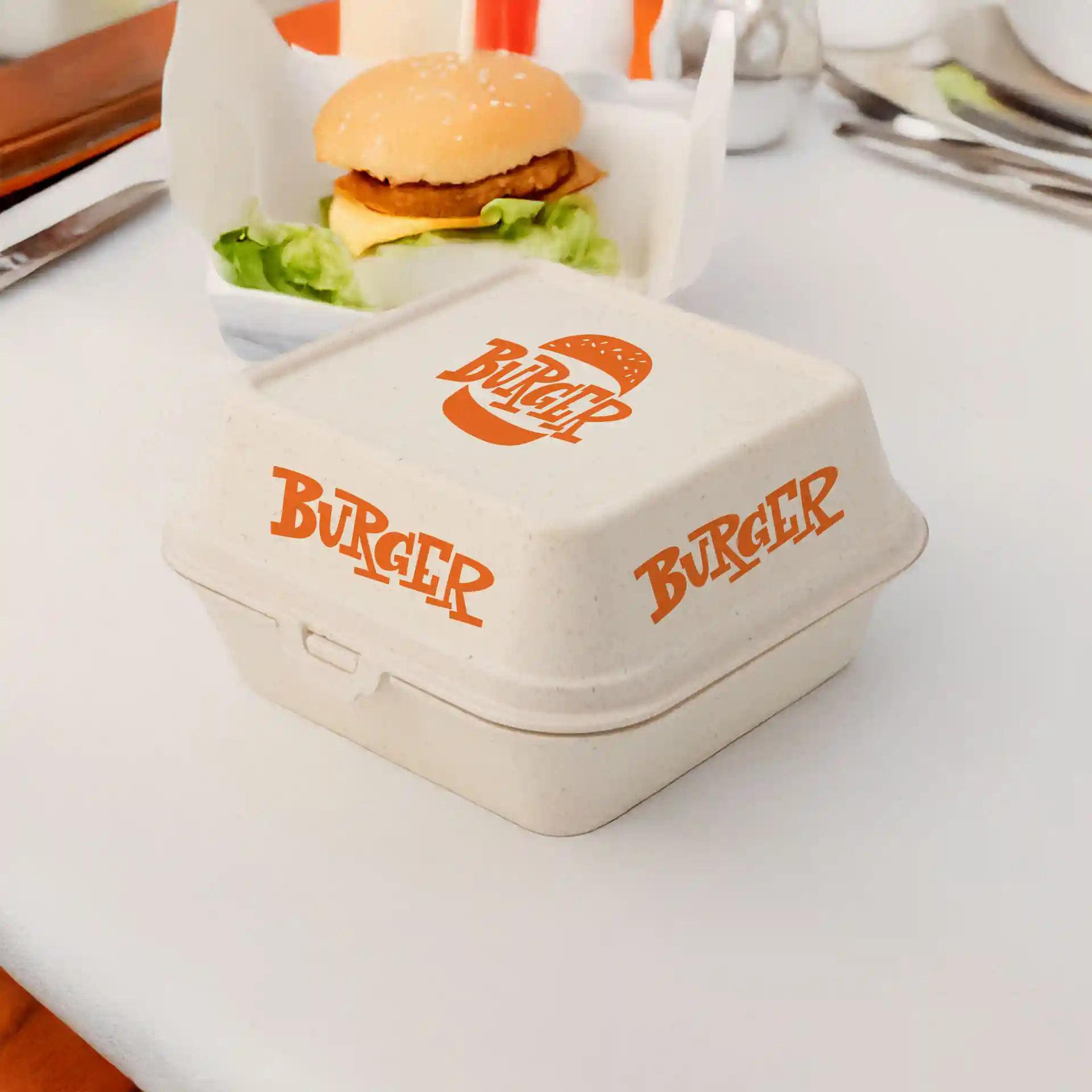 Boîte à burger rectangulaire crème blanche avec texte imprimé orange, placée sur une table à côté d'un burger et de laitue.
