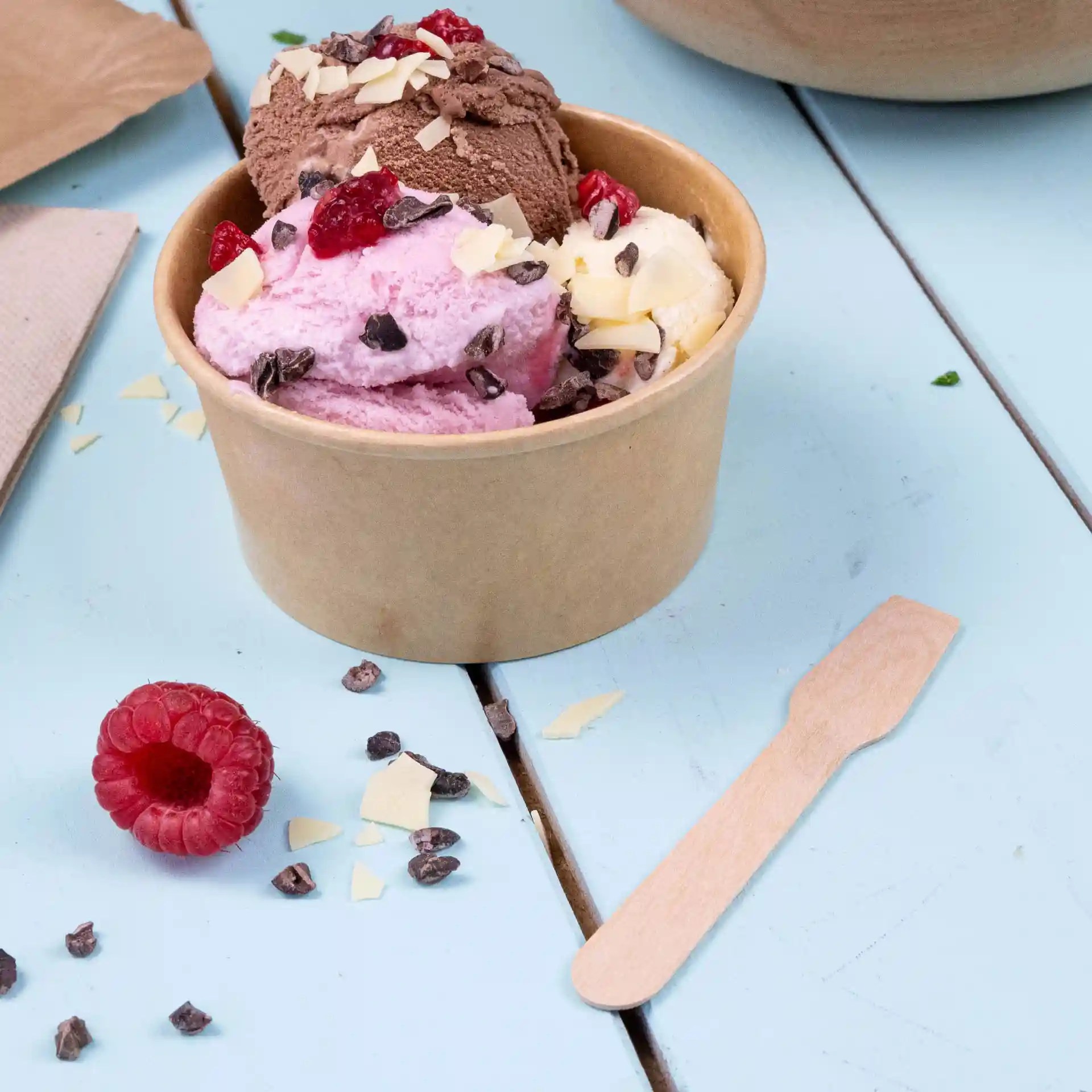 Becher mit drei Kugeln Eis in Pink, Braun und Weiß, mit Toppings, neben einem Holzlöffel und einer Himbeere.