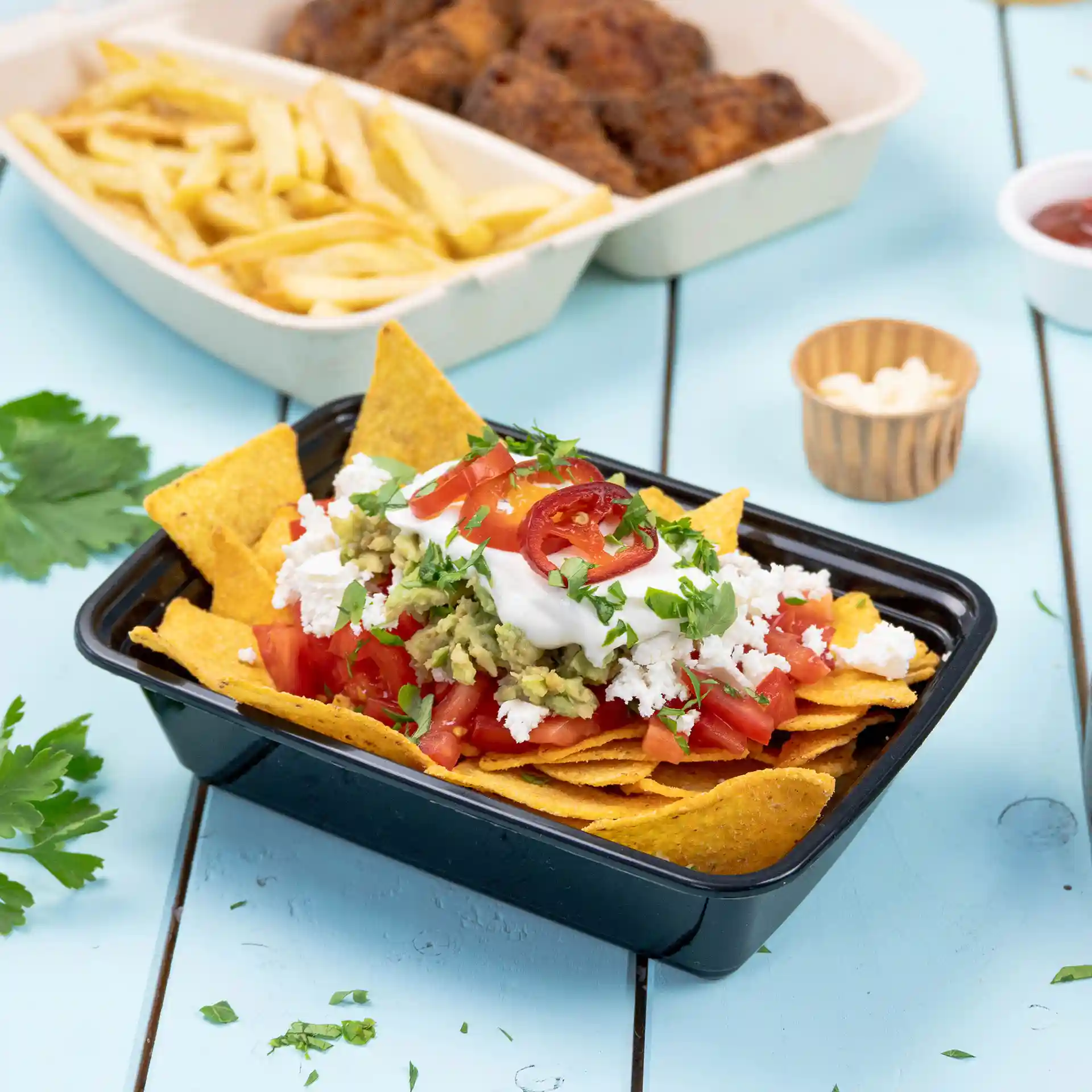 Schwarzer rechteckiger wiederverwendbarer Behälter gefüllt mit Nachos, belegt mit Tomaten, Guacamole, Sauerrahm und Koriander.