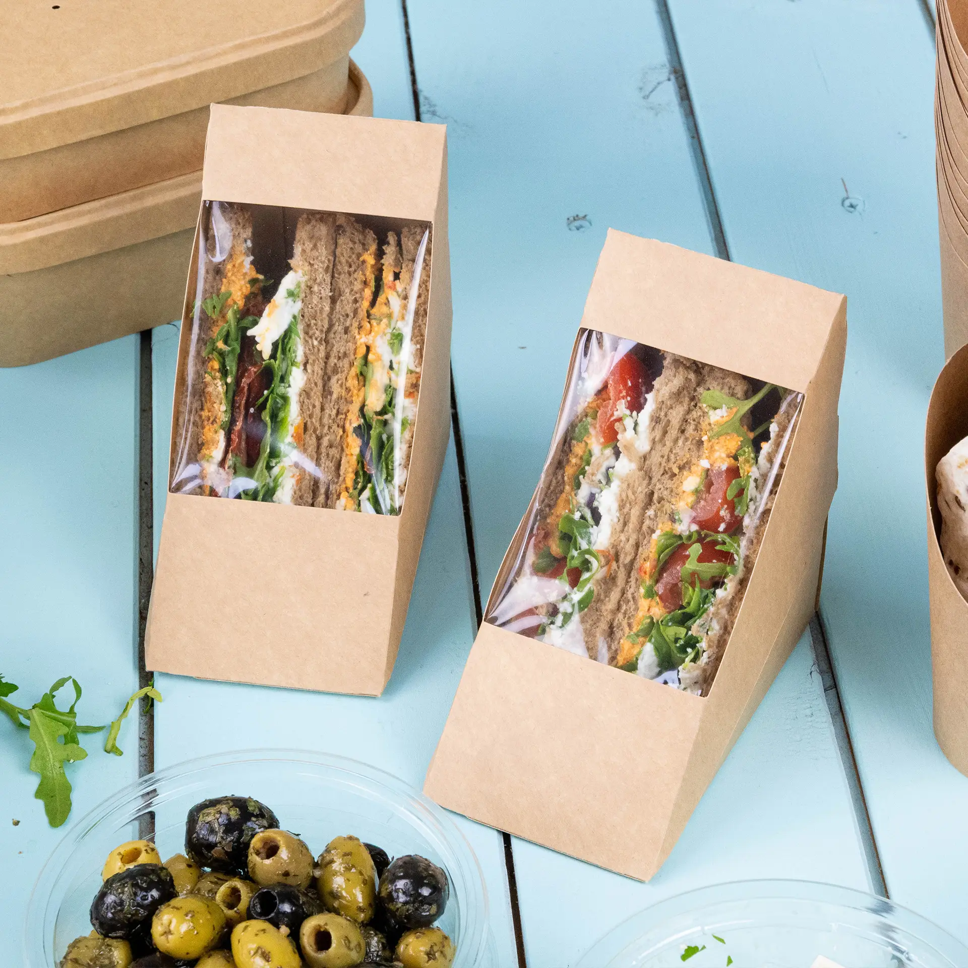 Due angoli di sandwich in cartone marrone con una finestra in PLA trasparente che mostra sandwich e panini farciti su una superficie blu, con olive in una ciotola nelle vicinanze.