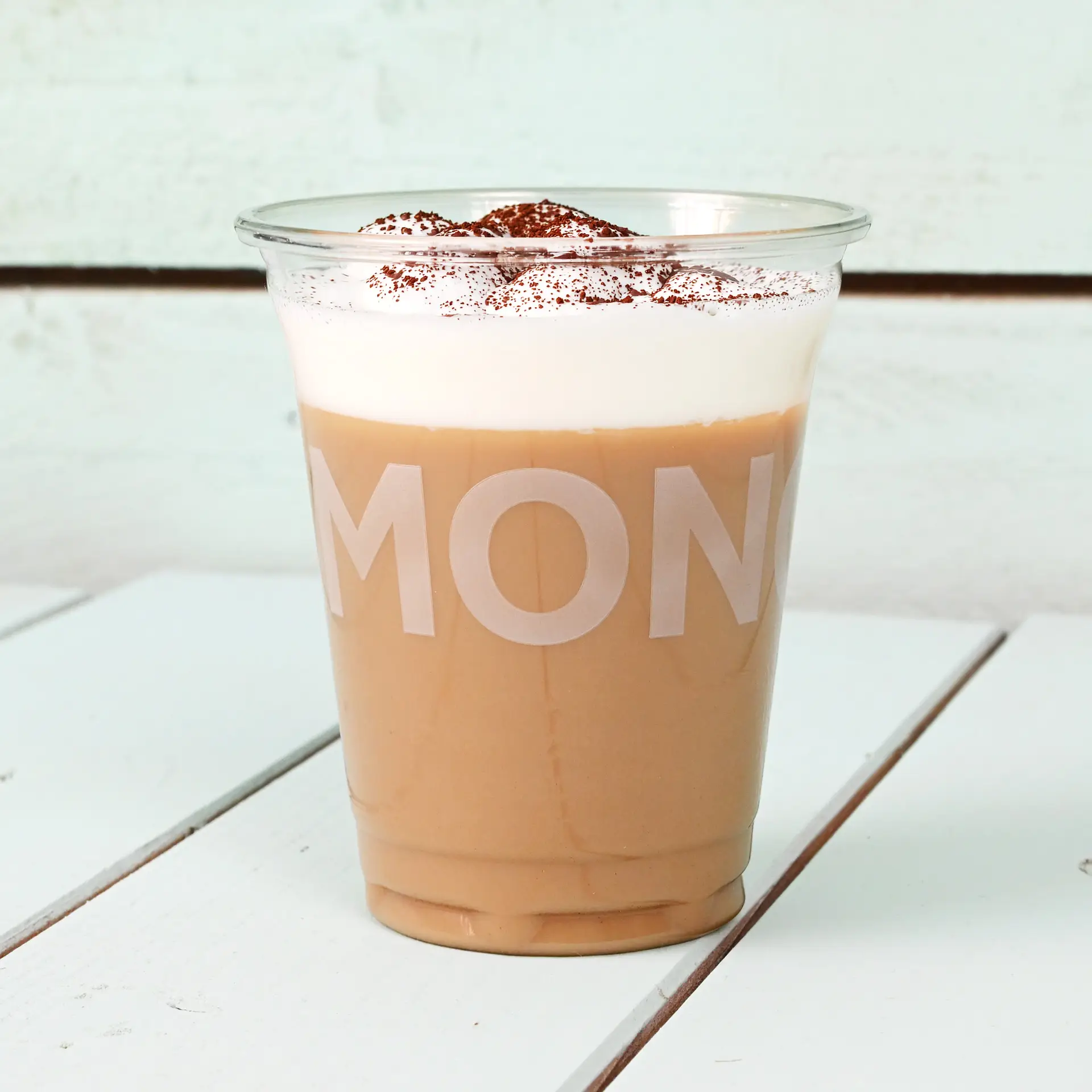 Bicchiere rPET trasparente ripieno di caffè ghiacciato, guarnito con panna montata e cacao, su un piano in legno.