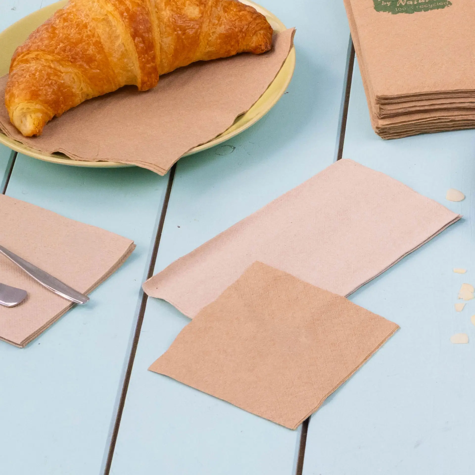 Immagine di un croissant su un piatto giallo con diversi tovaglioli da cocktail in carta riciclata marrone disposti su un tavolo blu.