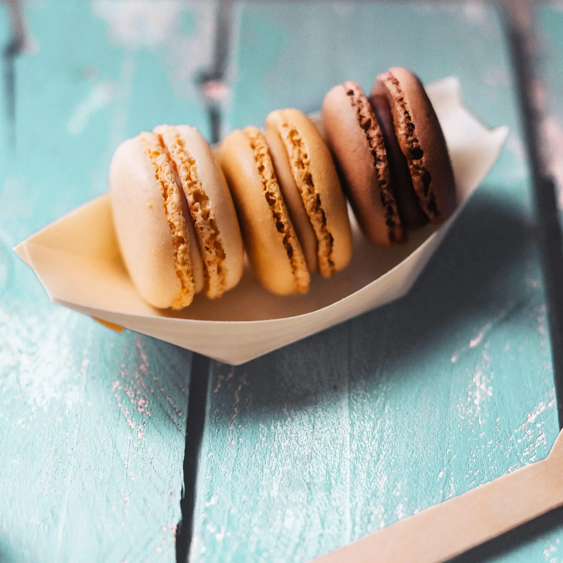 Vier Macarons in einem bootförmigen Halter, in beige, hellbraun, dunkelbraun und schokoladenfarben auf einer strukturierten Oberfläche.