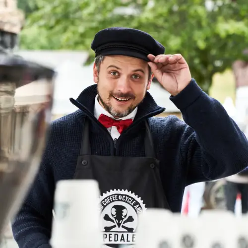Homme en casquette noire et nœud papillon, portant un tablier de marque, souriant et saluant les visiteurs à un stand de café.