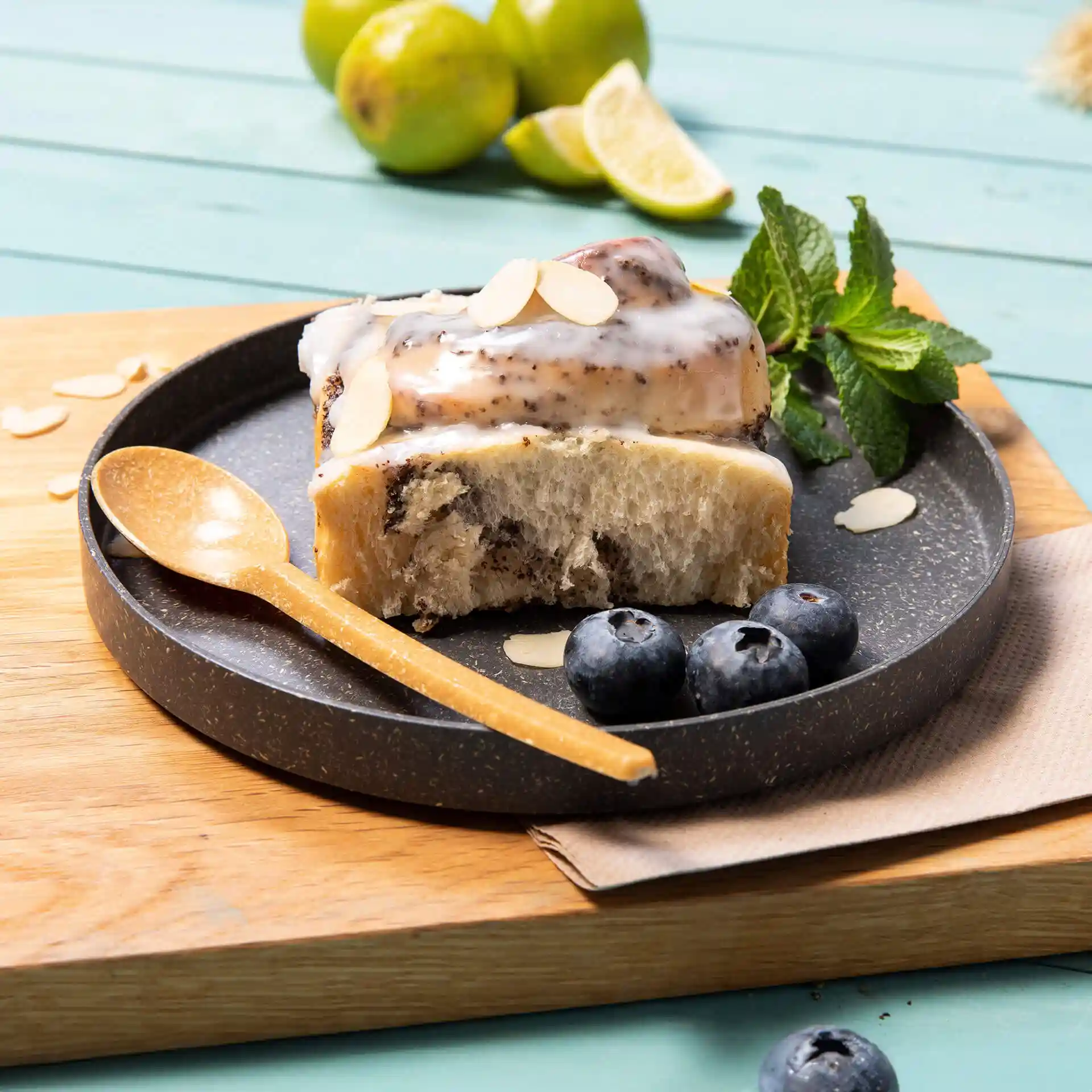 Stück Kuchen mit Zuckerguss und Mandeln auf einem grauen runden Teller, begleitet von Heidelbeeren und einem Holzlöffel.