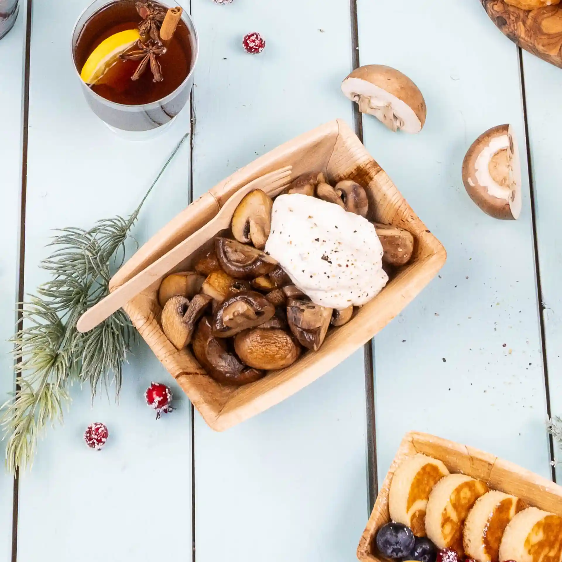 Bol rectangulaire en feuille de palmier avec des champignons sautés et une cuillerée de crème, accompagné d'une boisson et d'éléments décoratifs.