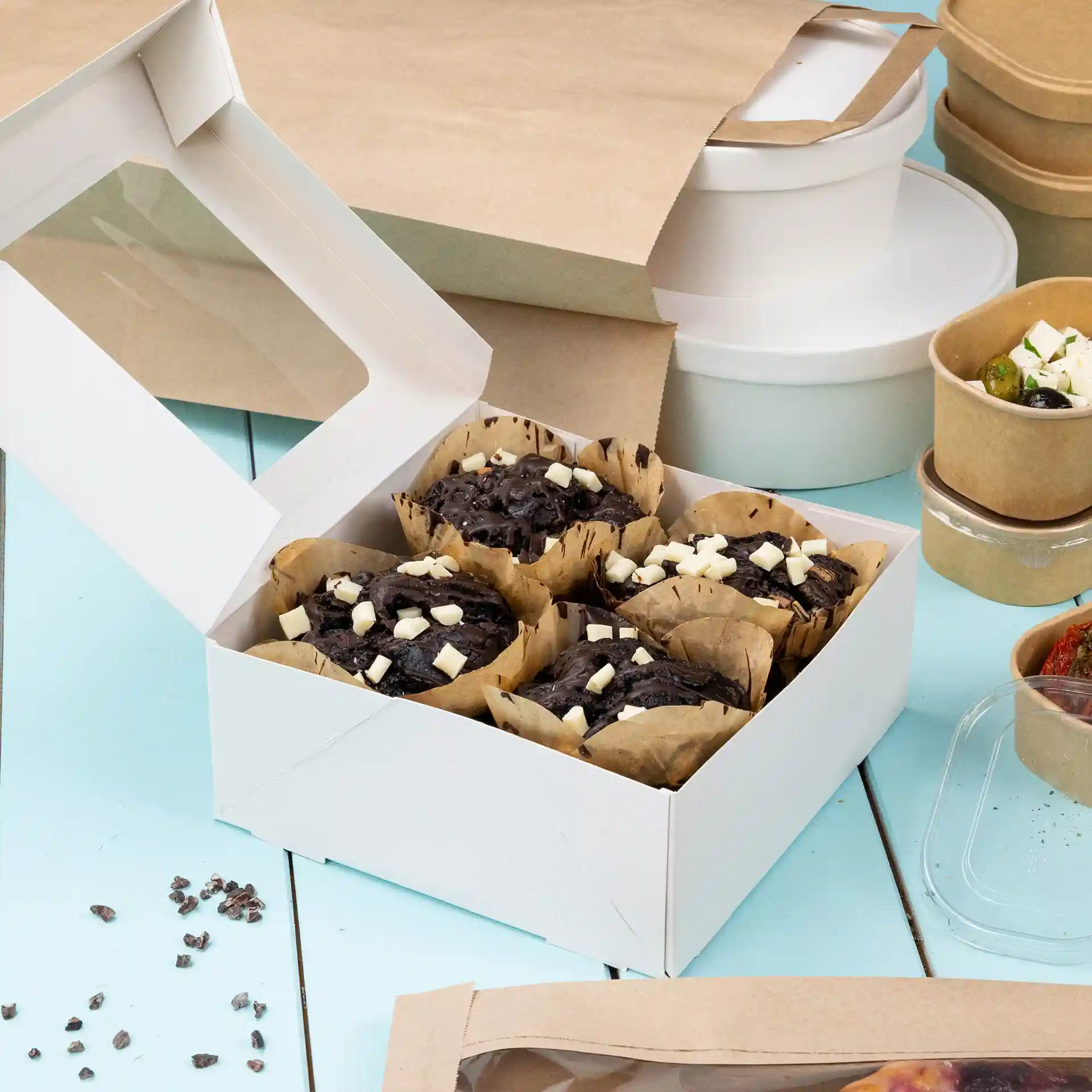 Weiße Pappschachtel mit klarem Fenster, gefüllt mit mehreren Schokoladenmuffins, die mit weißen Schokoladenstückchen belegt sind.