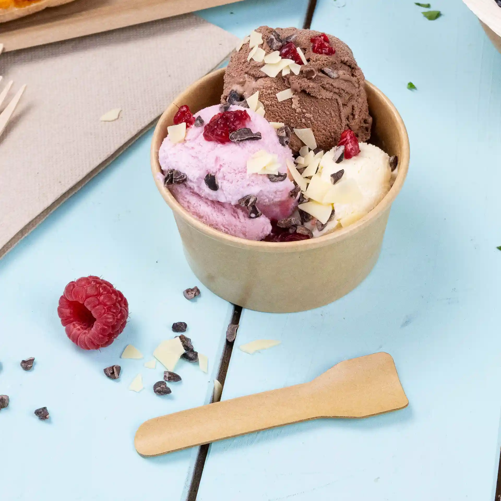 Becher mit drei Kugeln Eis in Pink, Braun und Weiß, mit Schokoladenstückchen und Himbeeren, auf einer blauen Oberfläche.