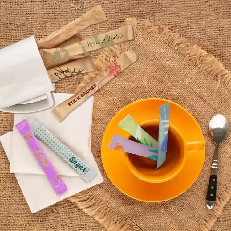 Sachets de sucre assortis placés dans une tasse de thé sur une surface en jute, avec une cuillère et une serviette à proximité.