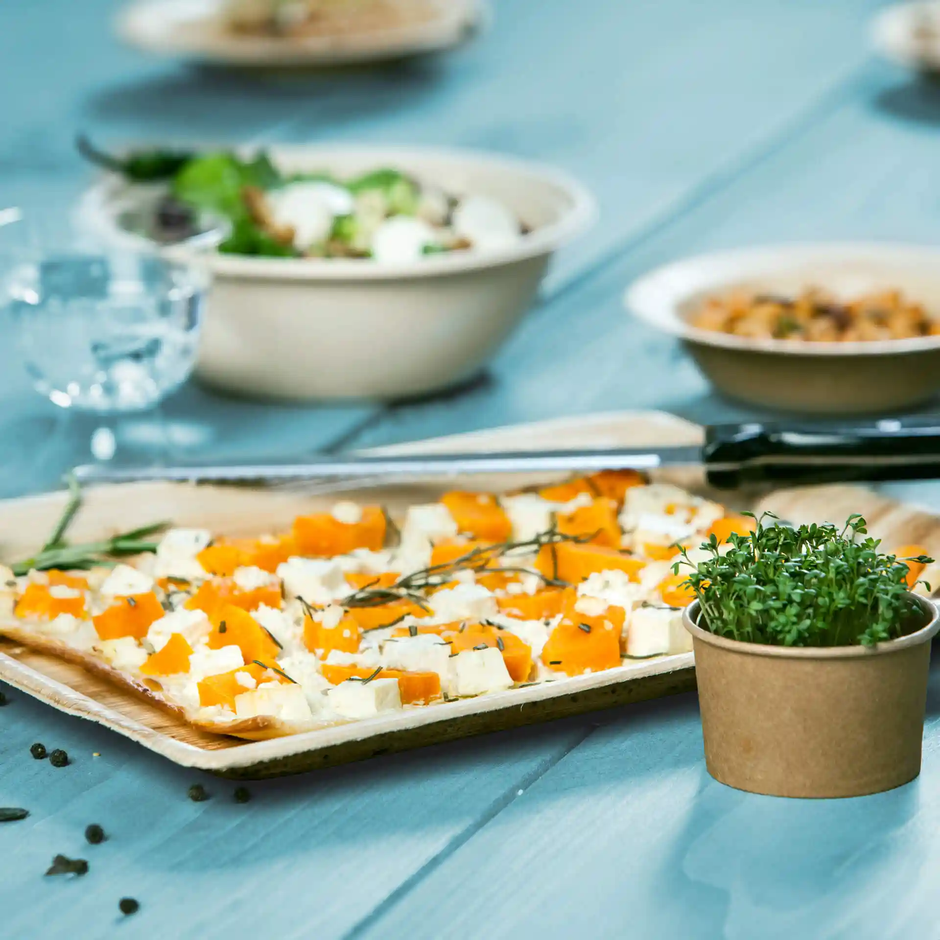 Vassoio rettangolare in foglia di palma con pezzi di cibo arancioni e bianchi, accompagnato da un piccolo vaso di microgreens su un tavolo blu.