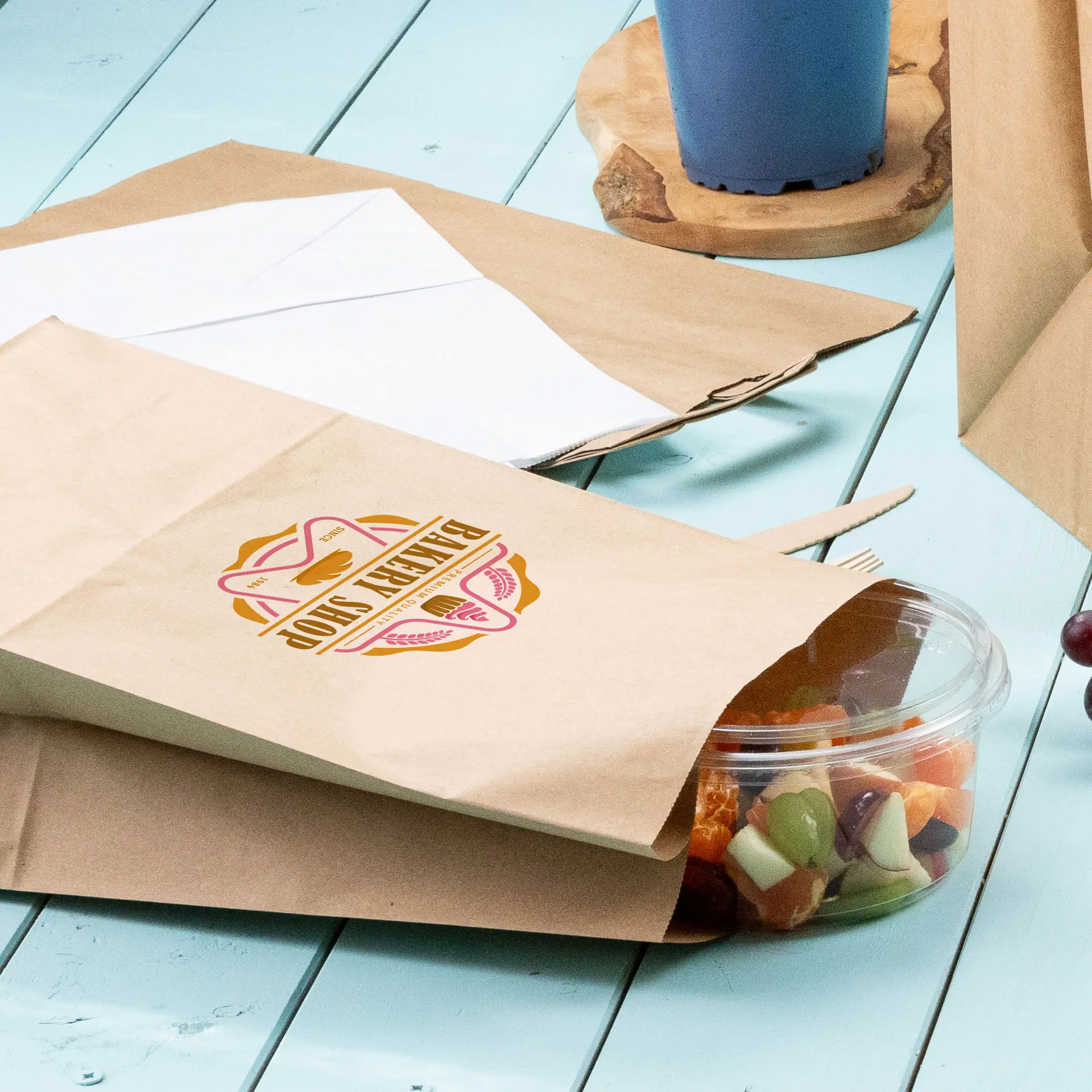 Borse di carta marroni stampate con un design circolare e un contenitore di plastica trasparente con frutta mista su una superficie blu.