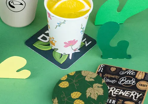 Stapel von Untersetzern auf einer Holzoberfläche; einige zeigen ein Logo mit einem Vogel, andere haben ein Design mit Bier und Text.