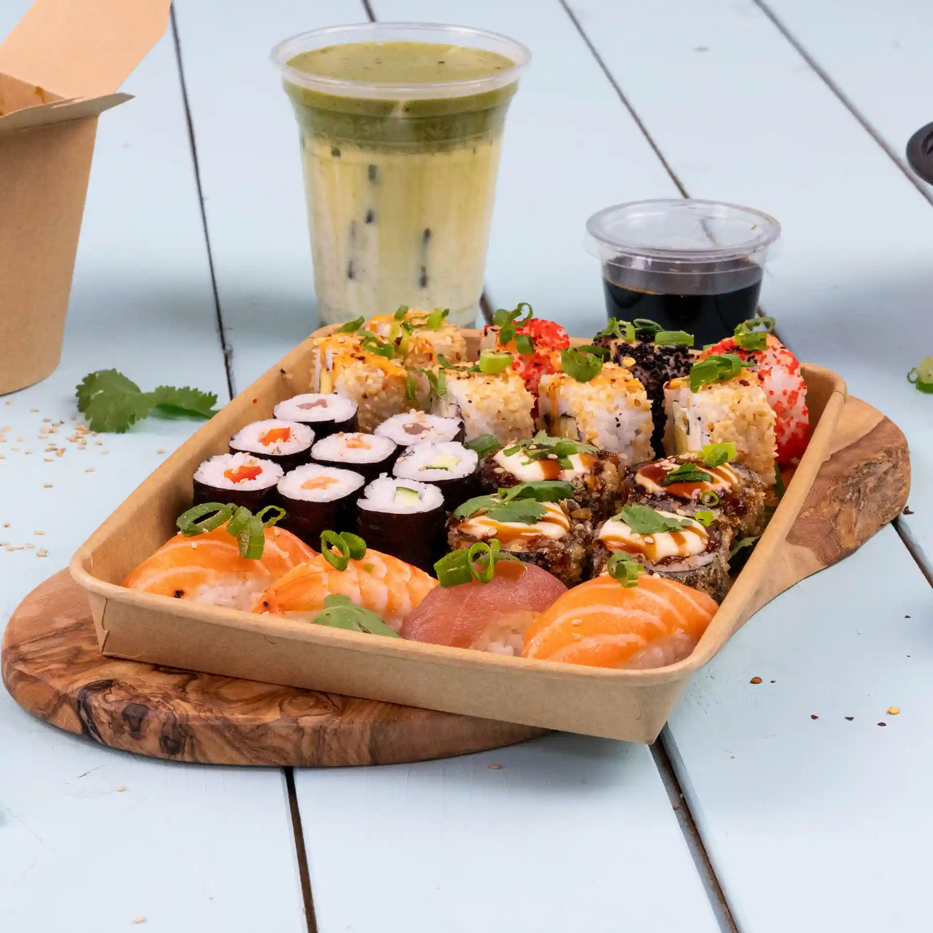 Tablett mit verschiedenen Sushi, darunter Rollen und Sashimi, serviert mit einem Getränk und Sojasauce auf einem Holzbrett.