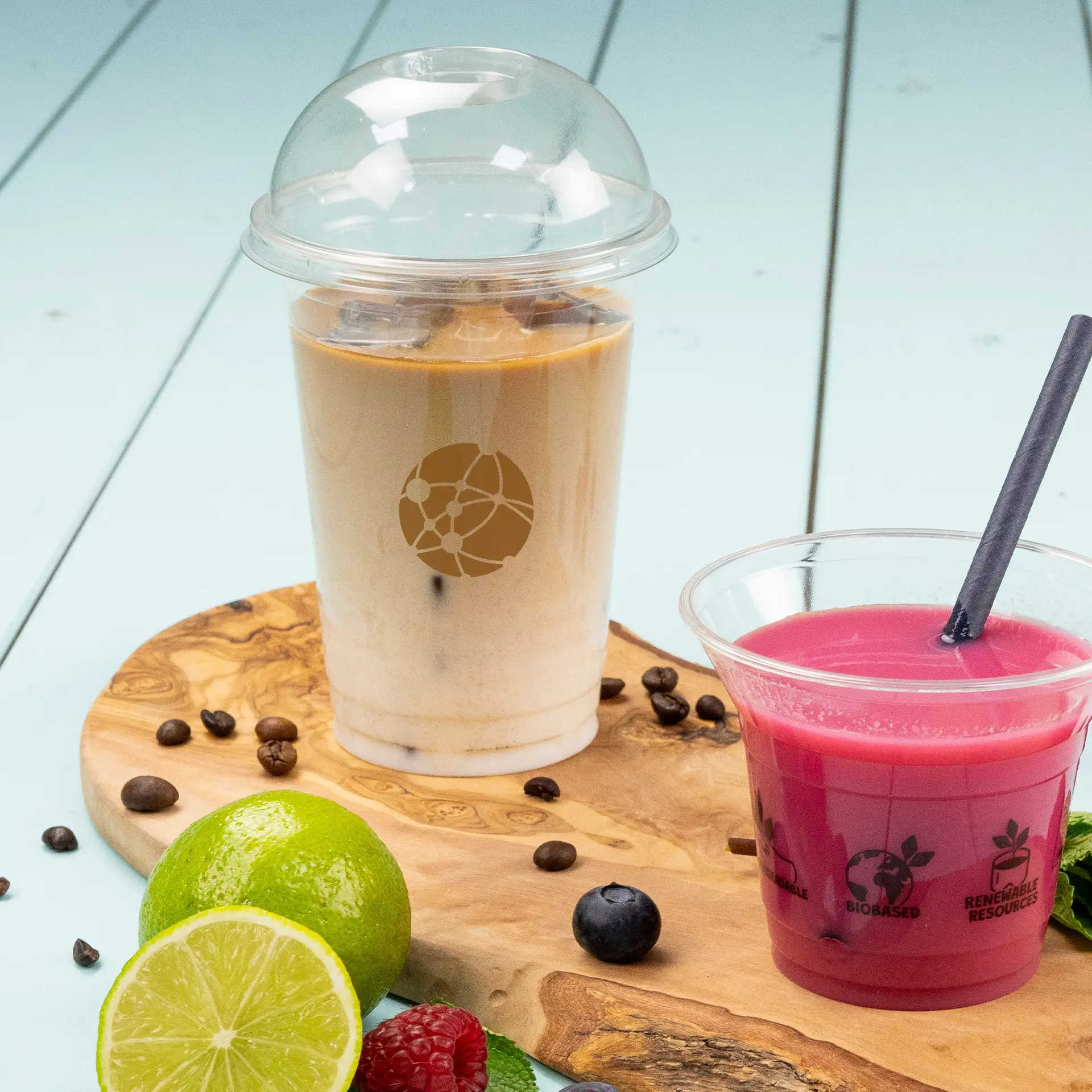 Un cup PLA trasparente riempito di caffè freddo accanto a un frullato rosa; lime fresco, bacche e chicchi di caffè su un tagliere di legno.