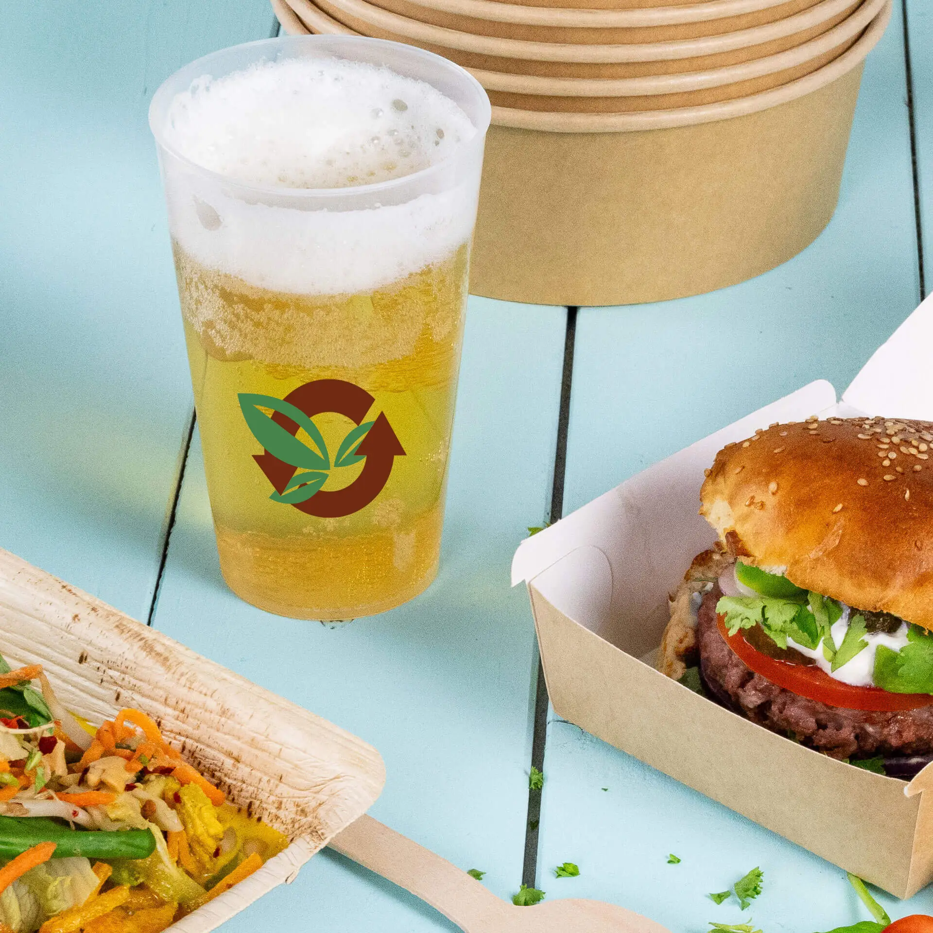 Gobelet réutilisable avec logo contenant de la bière, à côté d'un sandwich et d'une fourchette en bois sur une surface bleue.
