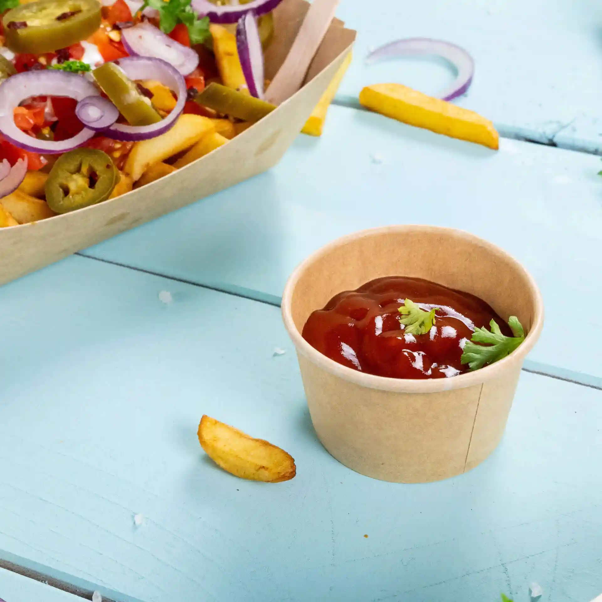 Cup in carta marrone con ketchup e un guarnizione di coriandolo, posato su una superficie azzurra con patatine vicino.