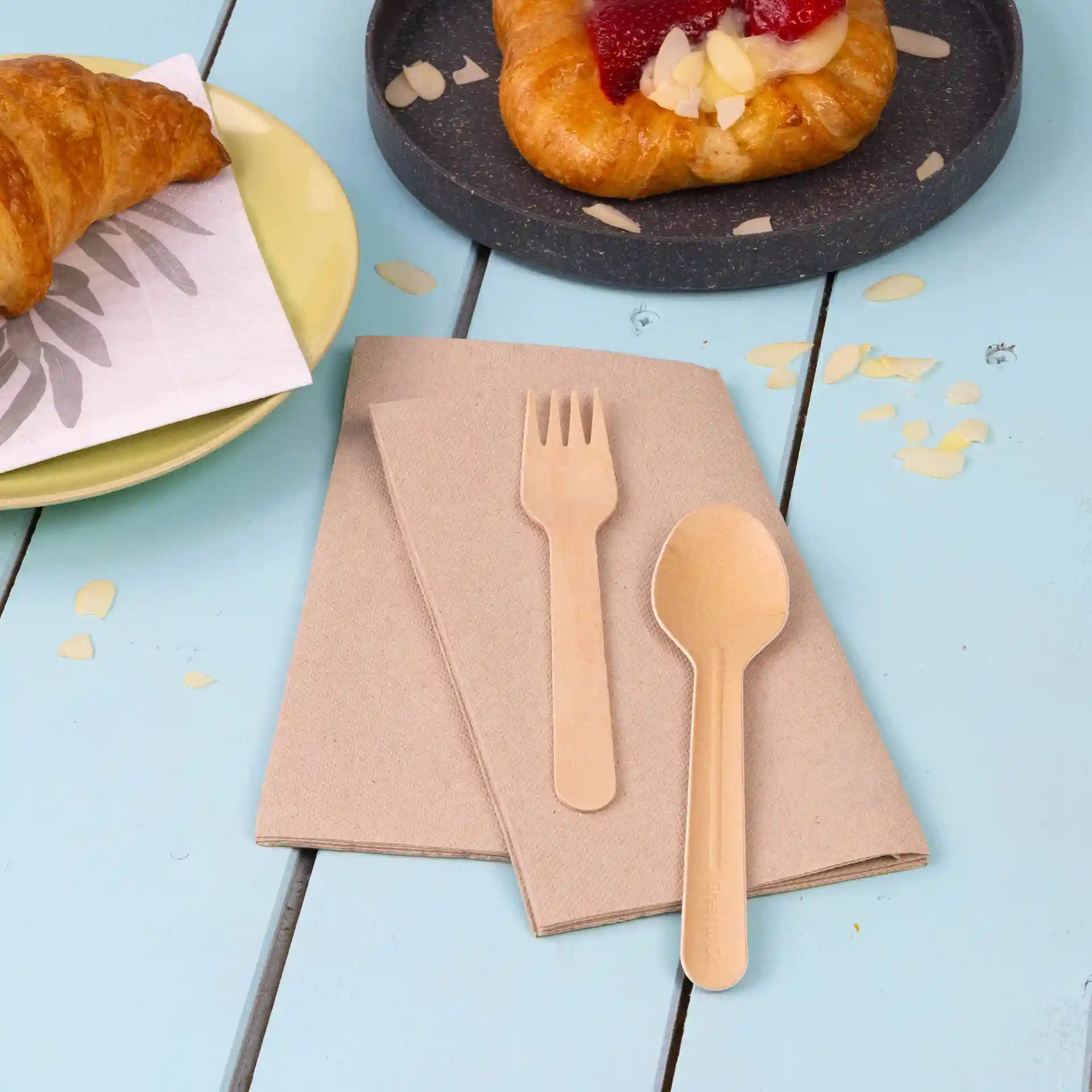 Ensemble de serviettes en papier recyclé non blanchi avec une fourchette et une cuillère en bois sur une table en bois bleue.