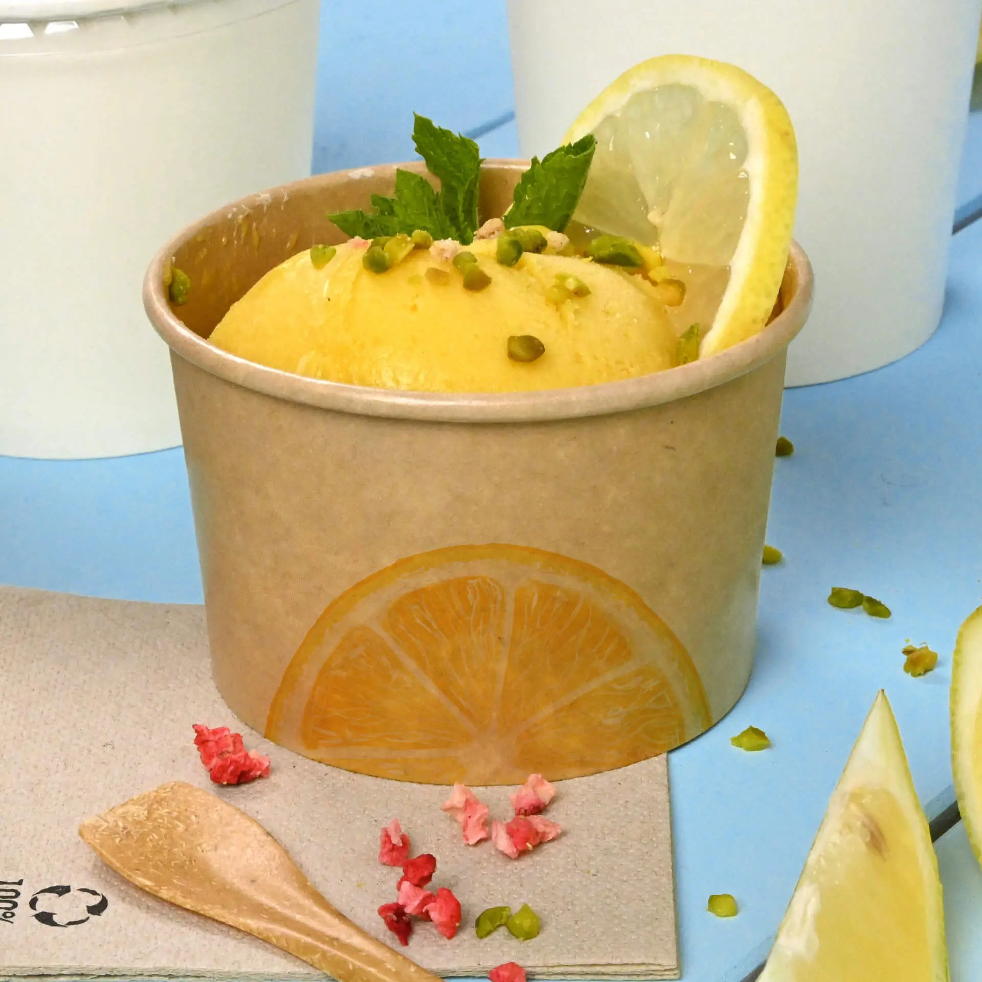 Brauner Becher mit Mango-Eis, Minzblättern und Zitronenscheibe, umgeben von Toppings und einem Holzlöffel.