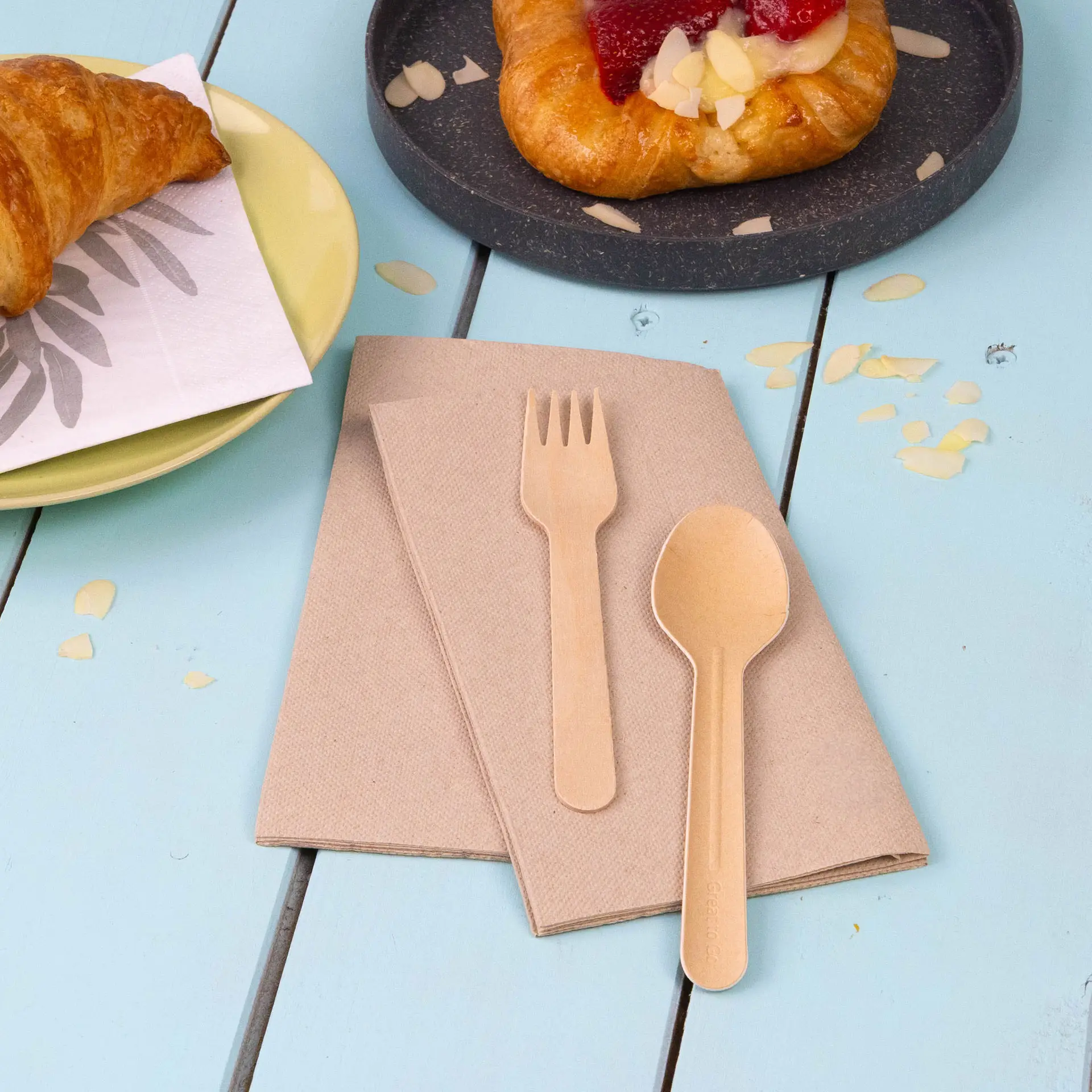 Zwei ungebleichte Recycling-Papierservietten mit einer Holzgabel und einem Holzlöffel auf einem blauen Tisch.